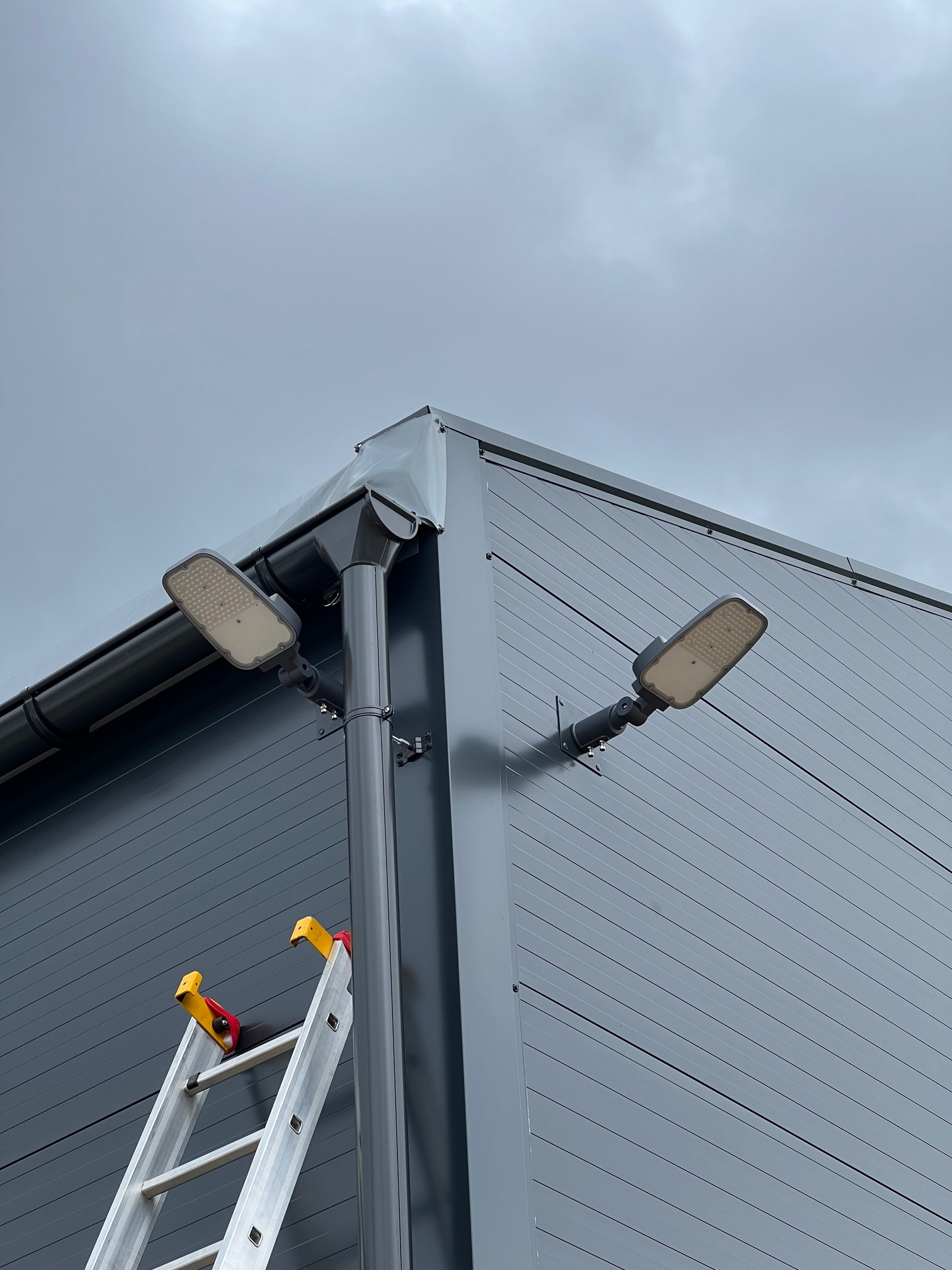 Dwa nowoczesne, prostokątne reflektory LED zamontowane na szarej, panelowej ścianie budynku, obok rynny spustowej; drabina aluminiowa oparta o ścianę, pochmurne niebo w tle.