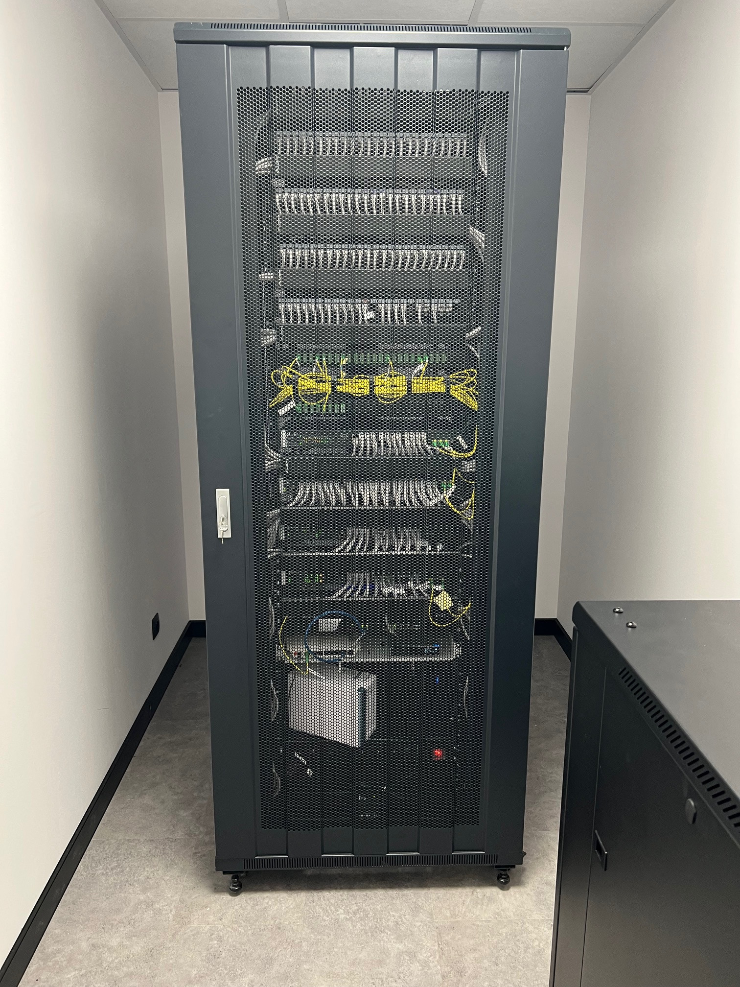 Szafa serwerowa z widocznymi kablami i urządzeniami, umieszczona w pomieszczeniu z białymi ścianami i szarą podłogą, obok widoczny fragment drugiej szafy.