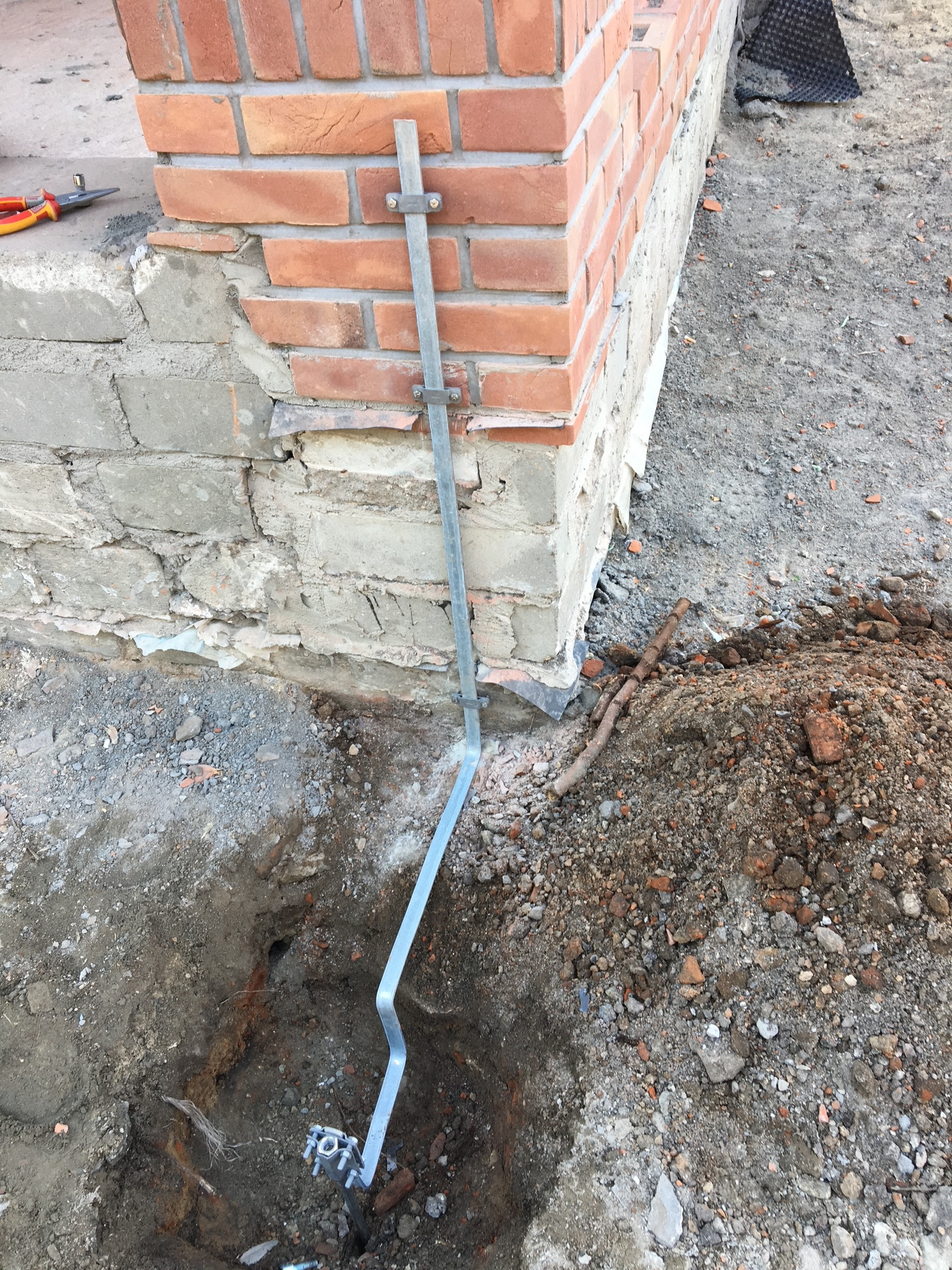 Uziemienie budynku w trakcie montażu, widoczna metalowa szyna przymocowana do fundamentu z cegły, częściowo zagłębiona w wykopie, obok leżą narzędzia.