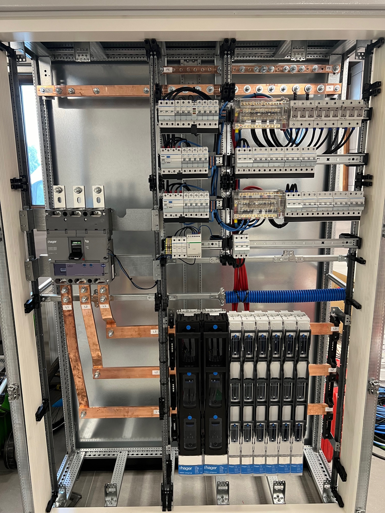 Wnętrze rozdzielnicy elektrycznej z widocznymi elementami: wyłącznik główny Hager, szyny miedziane oznaczone L1, L2, L3, zabezpieczenia modułowe, przewody w peszlu, szyny DIN, listwy zaciskowe.