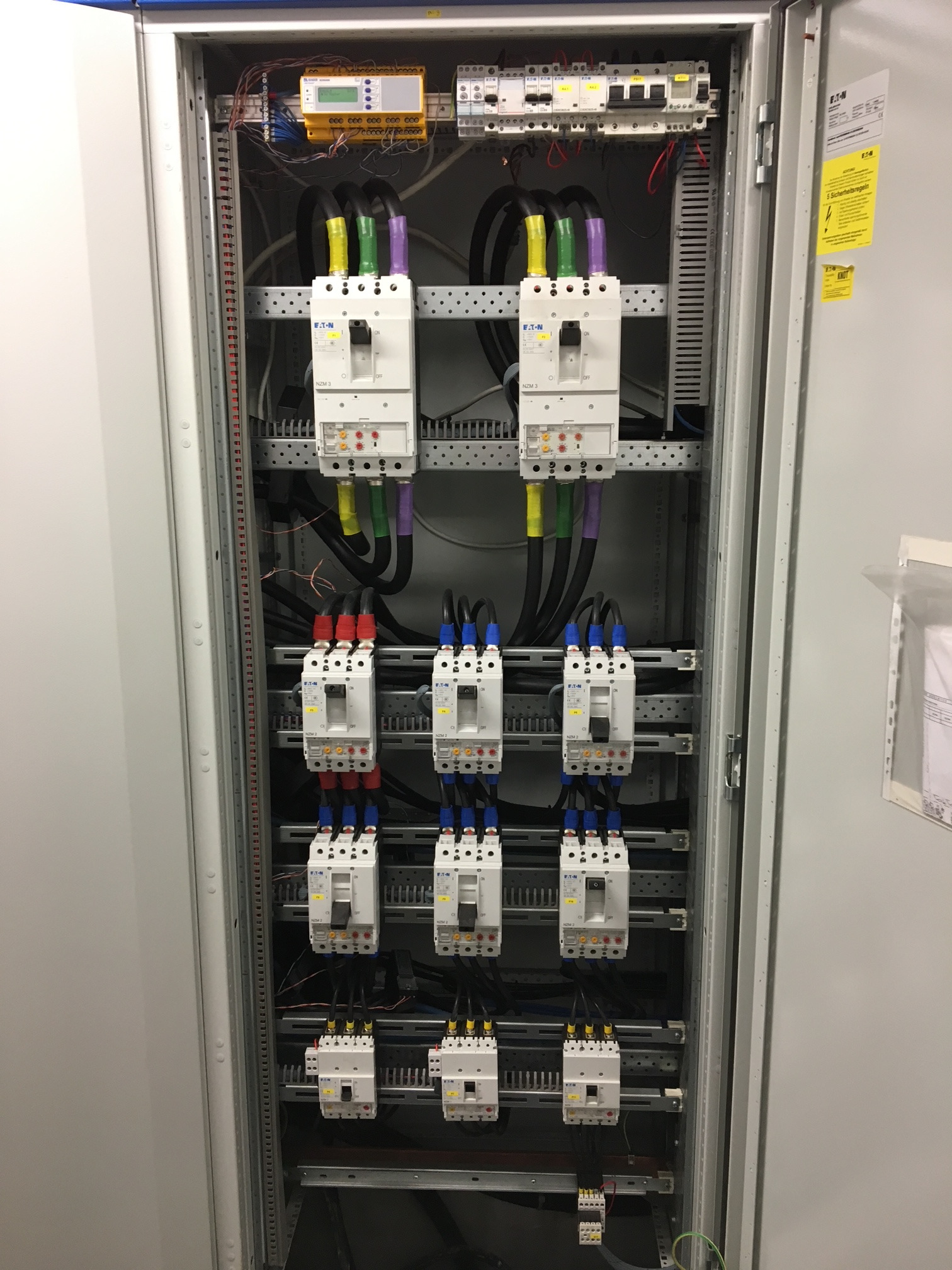 Rozdzielnia elektryczna z zamontowanymi bezpiecznikami i okablowaniem, widoczne przewody w kolorach czarnym, żółtym, zielonym i fioletowym, na górze żółty moduł sterujący, obok naklejka...