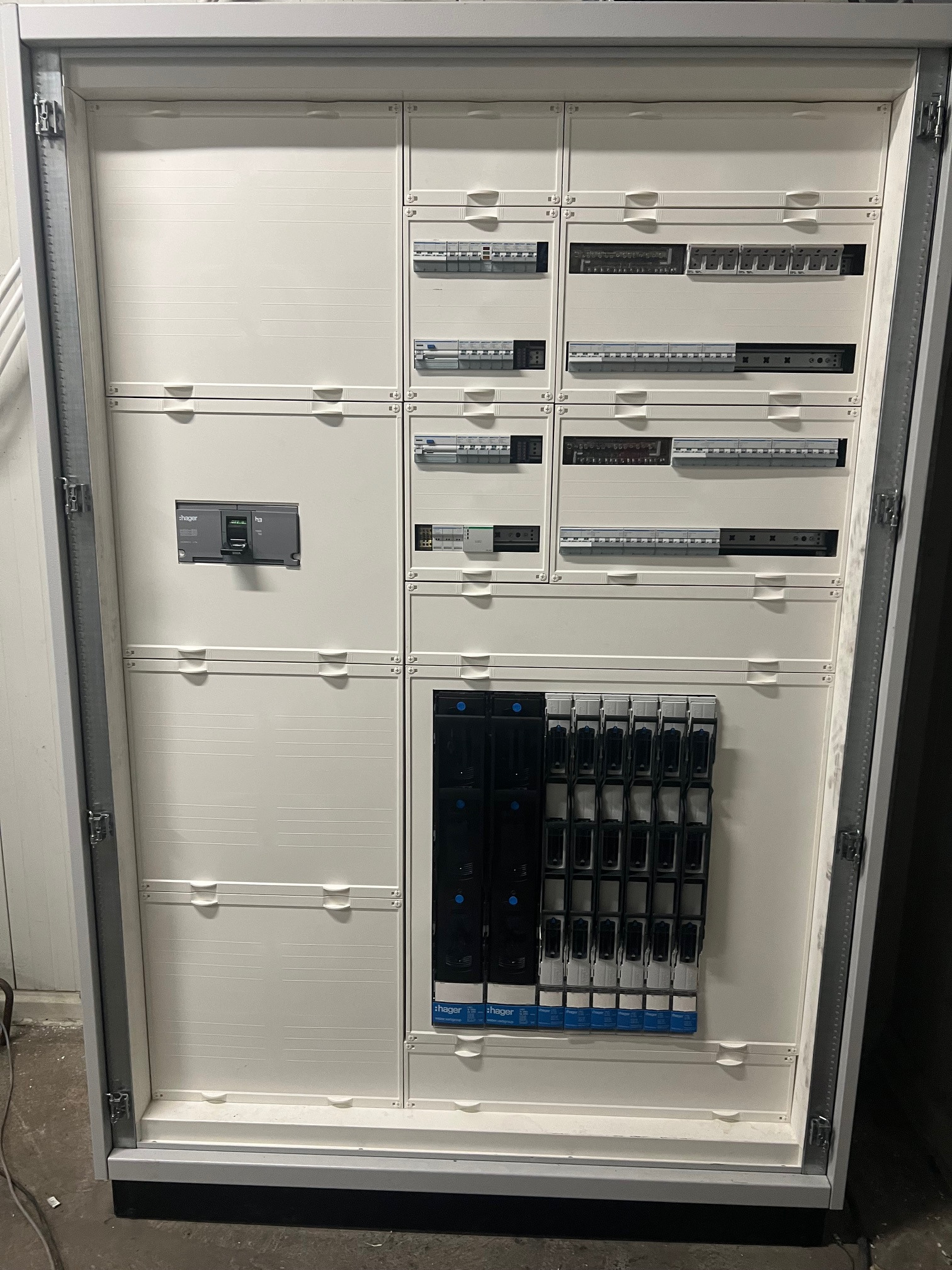 Otwarta, biała szafa rozdzielcza z zainstalowanymi modułowymi wyłącznikami i zabezpieczeniami firmy Hager, widoczna na tle ściany.