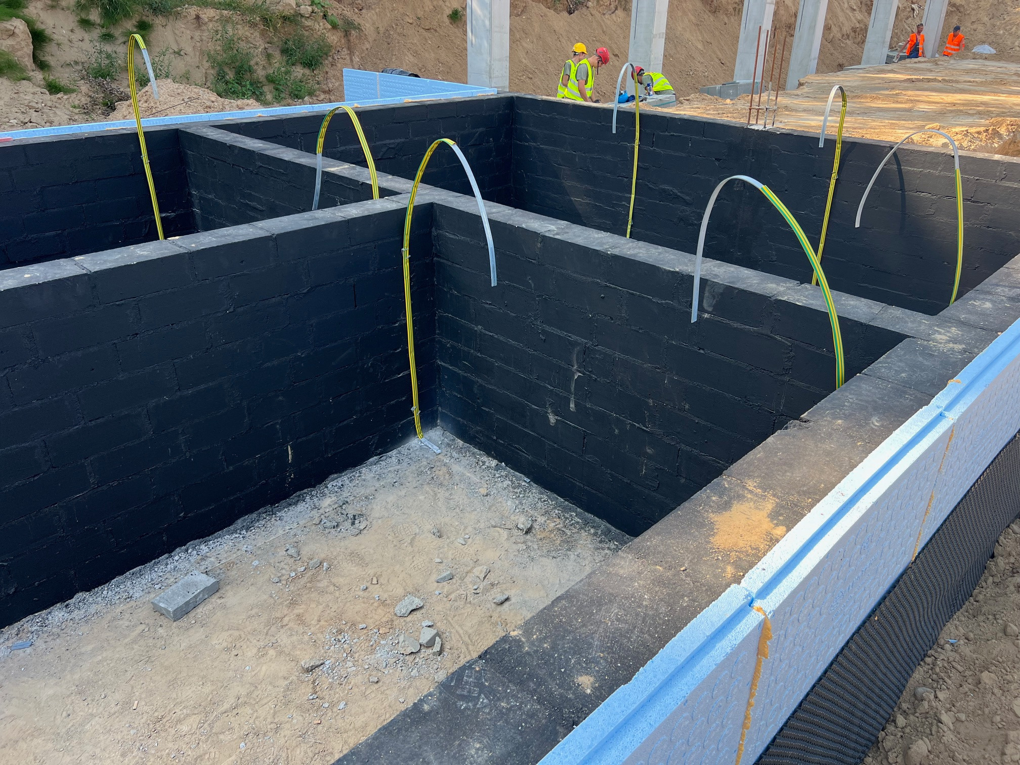 Fundamenty budynku z izolacją termiczną, czarną hydroizolacją i wyprowadzonymi przewodami elektrycznymi, w tle pracownicy w kamizelkach odblaskowych.