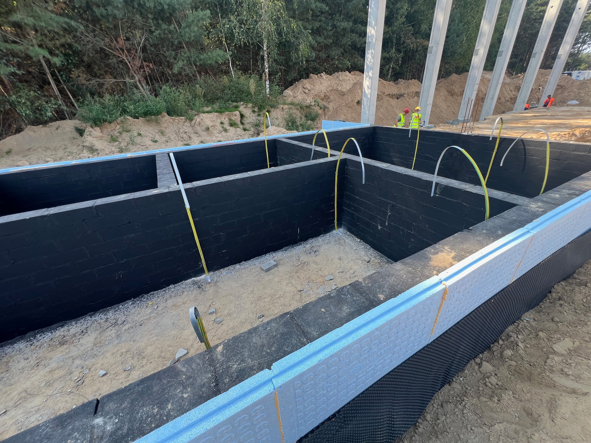 Fundamenty budynku z izolacją termiczną, czarną hydroizolacją i wystającymi przewodami elektrycznymi, widoczni pracownicy w oddali.