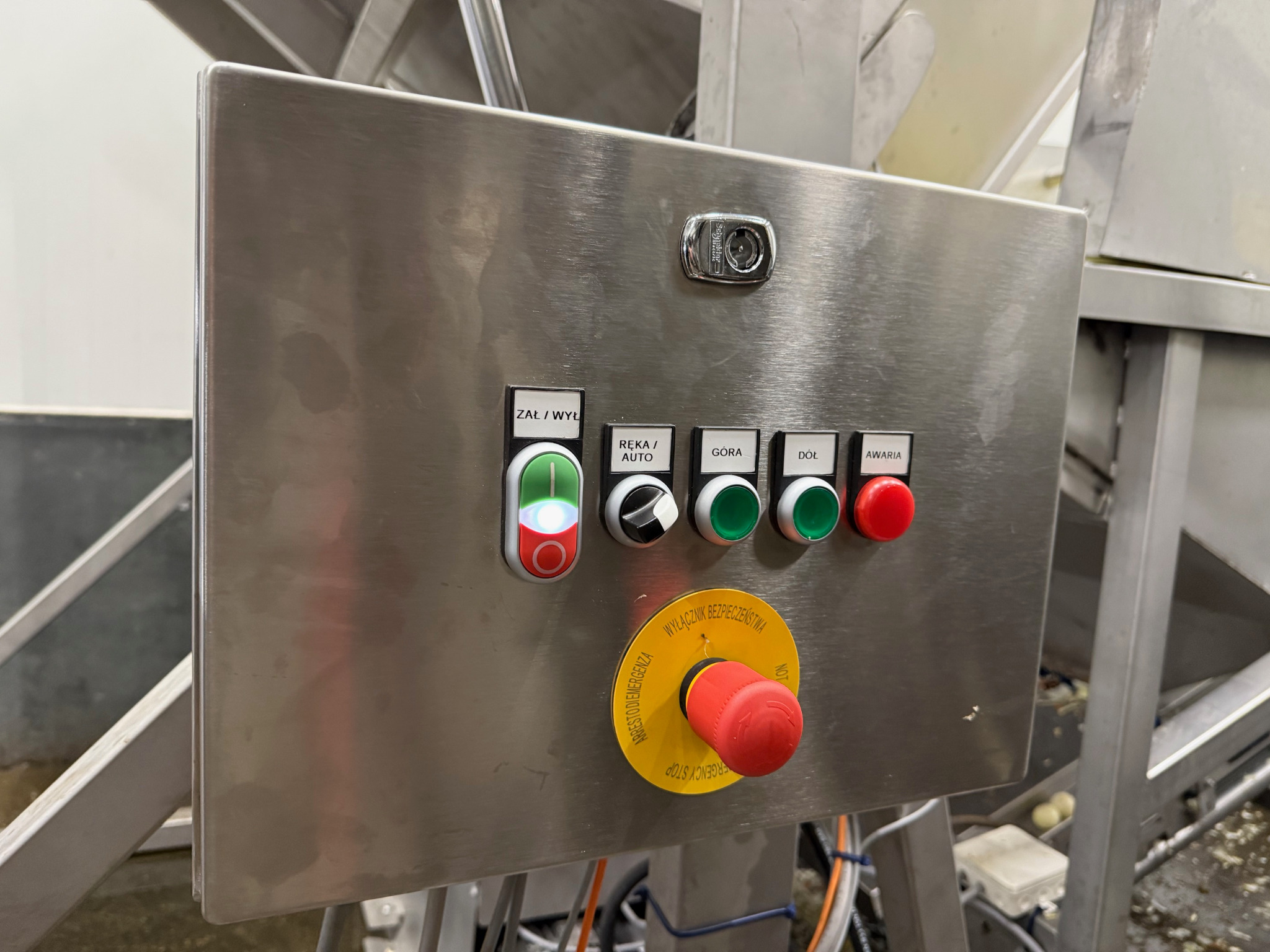 Panel sterowania ze stali nierdzewnej z przyciskami (włącz/wyłącz, ręka/auto, góra, dół, awaria) oraz wyłącznikiem bezpieczeństwa z żółtą obwódką, zamontowany na maszynie przemysłowej.