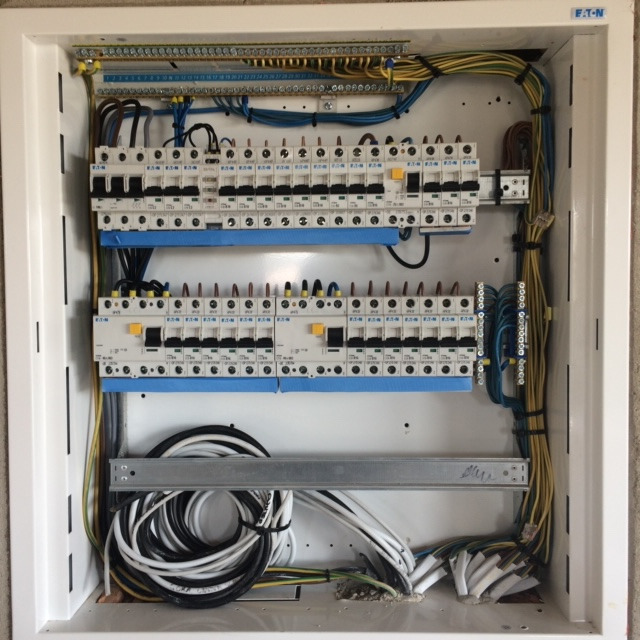 Rozdzielnica elektryczna marki Eaton, z widocznymi bezpiecznikami, listwami zaciskowymi i uporządkowanymi przewodami w kolorach: żółtym, niebieskim, czarnym i białym, umieszczona w białej obudowie.