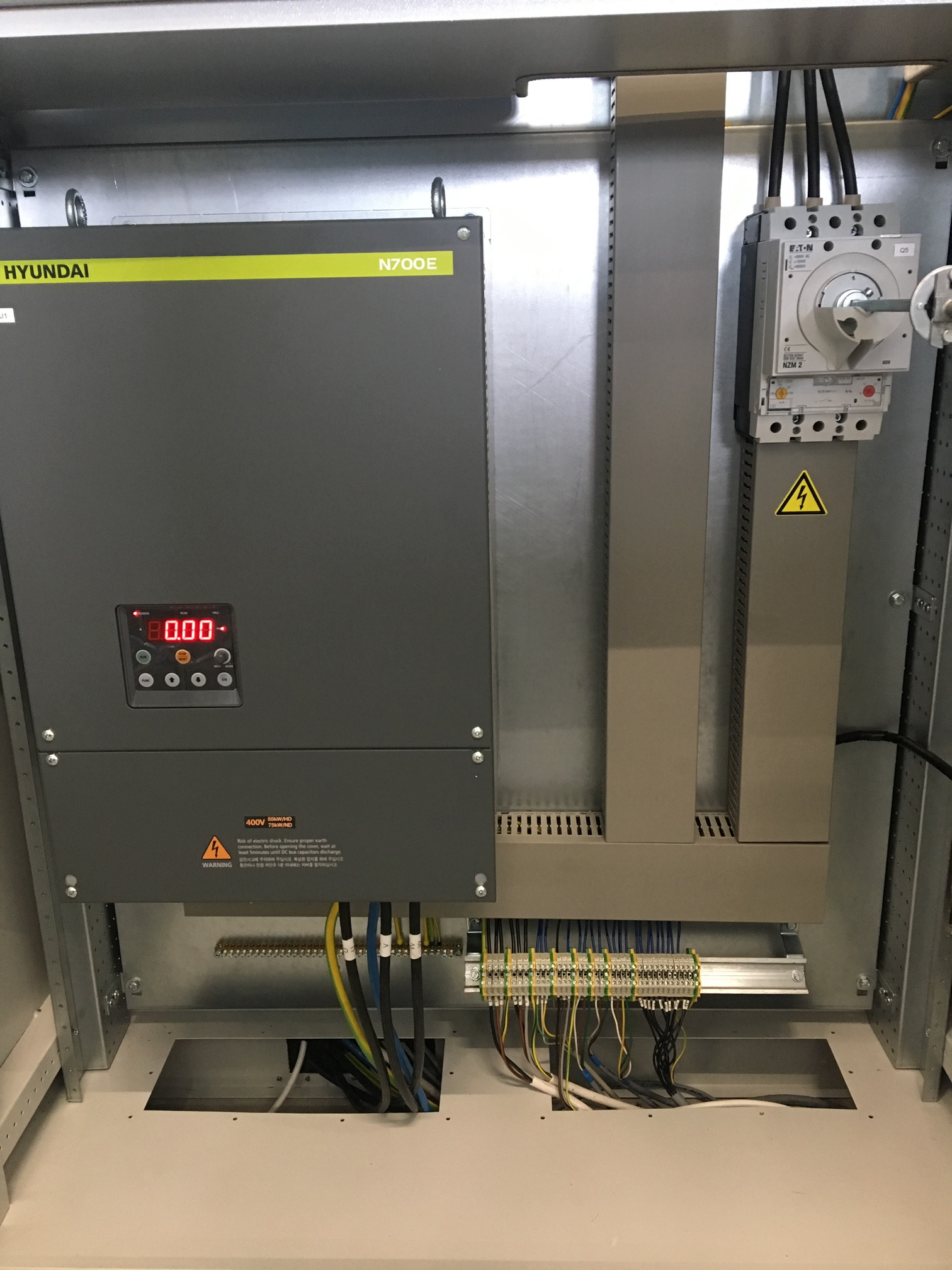 Wnętrze szafy sterowniczej z zamontowanym falownikiem Hyundai N700E, wyłącznikiem Eaton NZM2 oraz listwą zaciskową z podłączonymi przewodami. Widoczne oznaczenia ostrzegawcze o ryzyku porażenia...