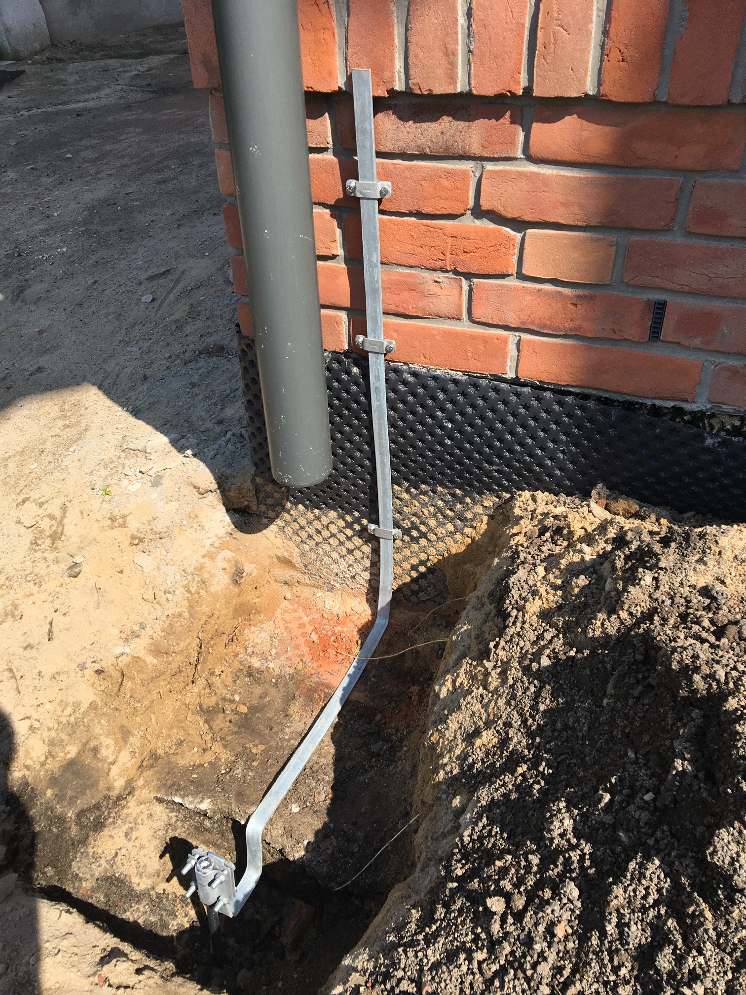 Uziemienie budynku w wykopie, widoczna metalowa szyna z mocowaniem przy ścianie z cegły, rura spustowa i czarna folia izolacyjna.