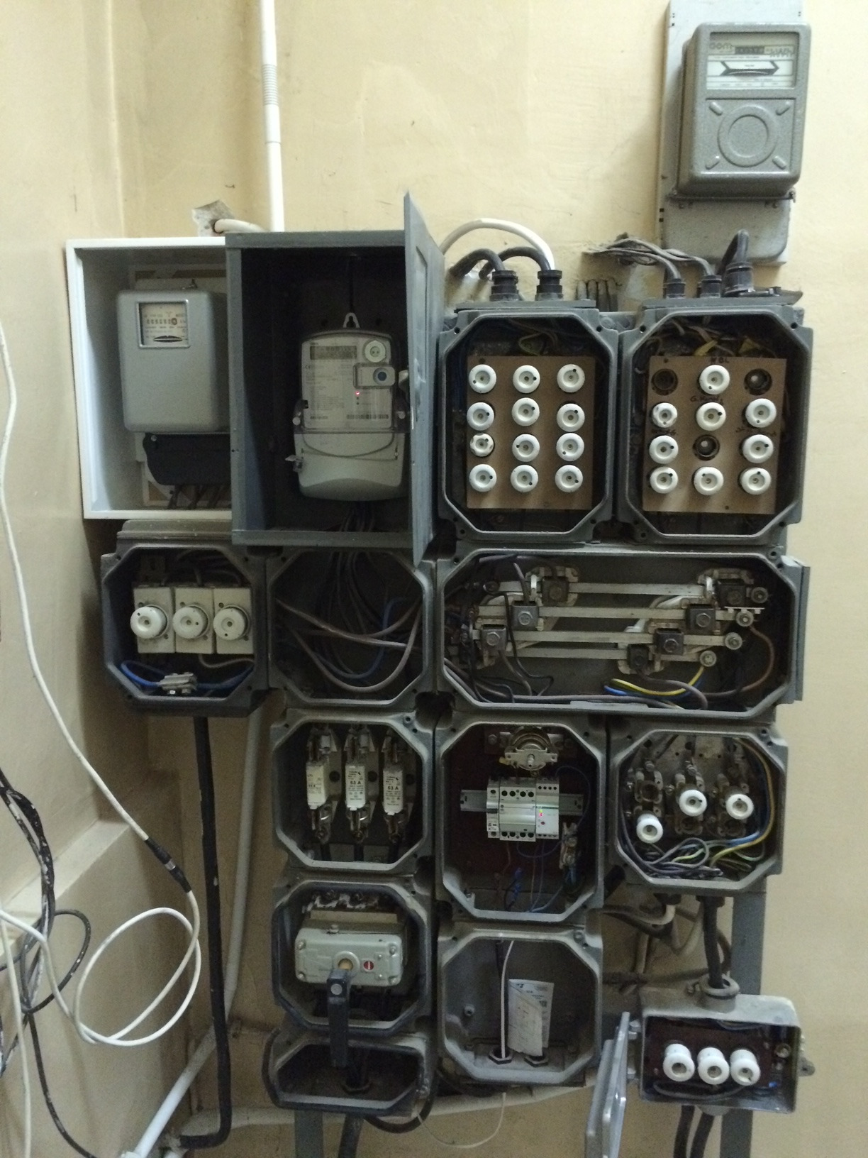 Rozdzielnia elektryczna z otwartymi obudowami, widoczne bezpieczniki topikowe, liczniki energii elektrycznej oraz okablowanie w starym budynku.