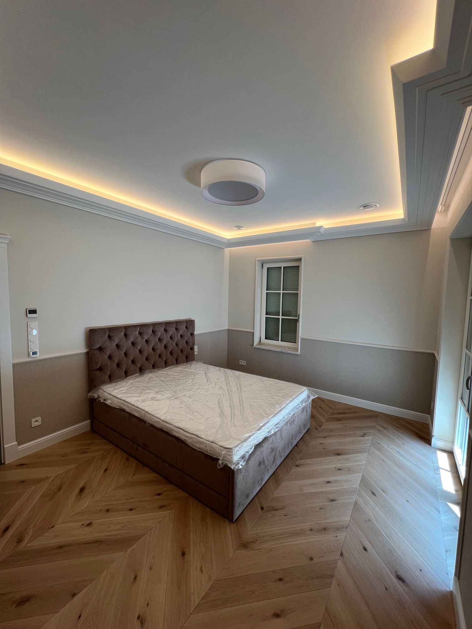 Sypialnia z łóżkiem w brązowej tapicerce, parkietem w jodełkę, oświetleniem LED w suficie i panelu sterowania na ścianie.