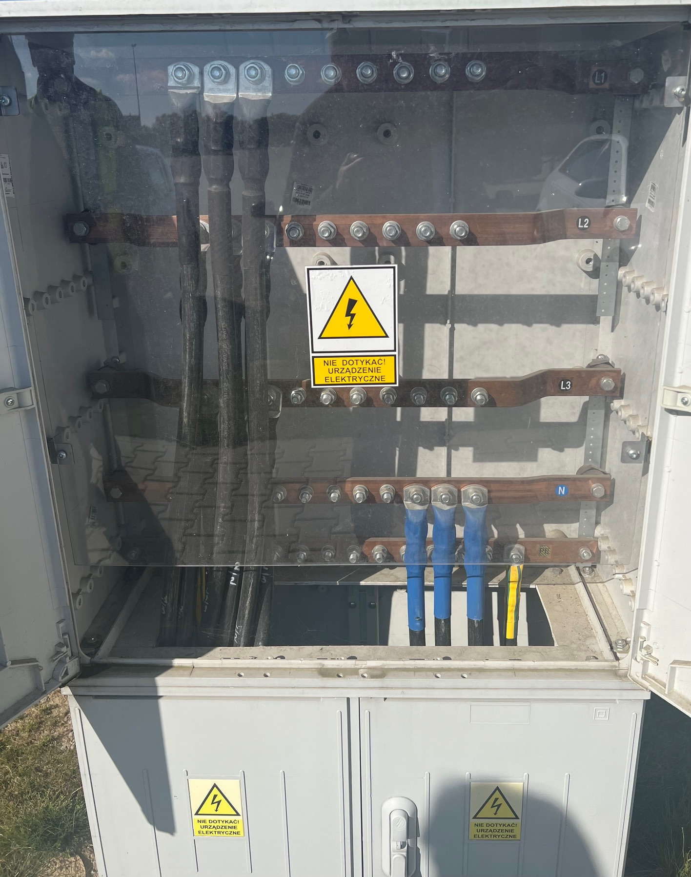 Otwarta skrzynka elektryczna z widocznymi przewodami, szynami zbiorczymi oznaczonymi L1, L2, L3, N oraz znakiem ostrzegawczym 'Nie dotykać urządzenia elektryczne'.