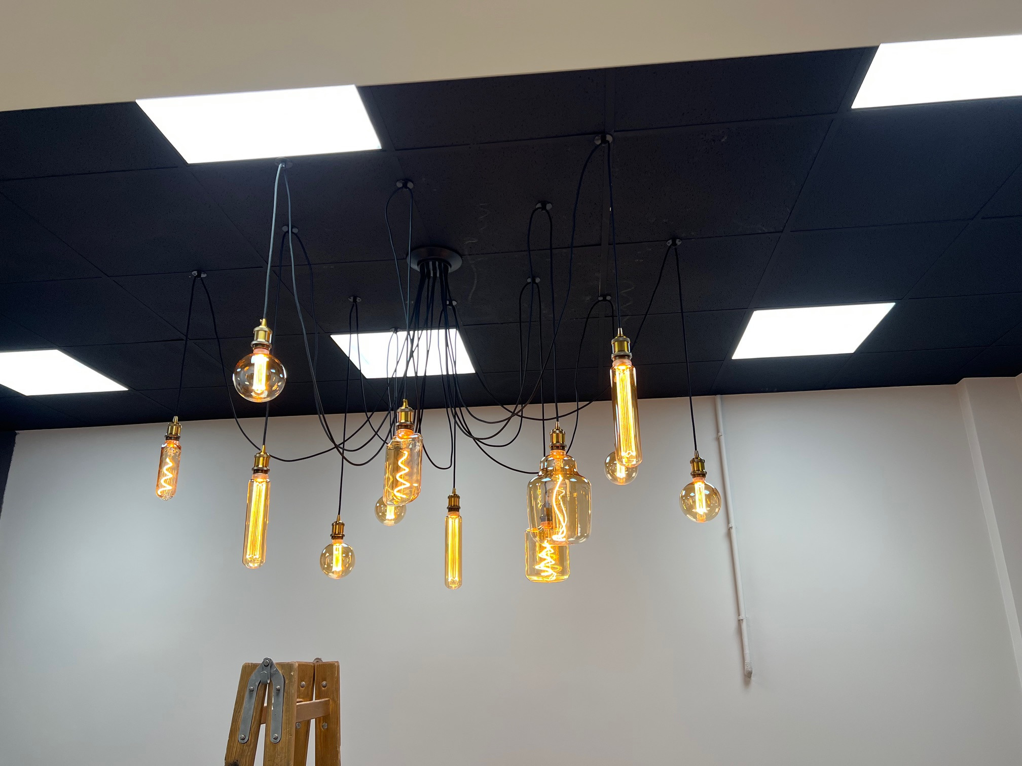 Dekoracyjna lampa wisząca z żarówkami Edisona o różnym kształcie, zamontowana na czarnym suficie z panelami oświetleniowymi, widoczny fragment drabiny malarskiej.