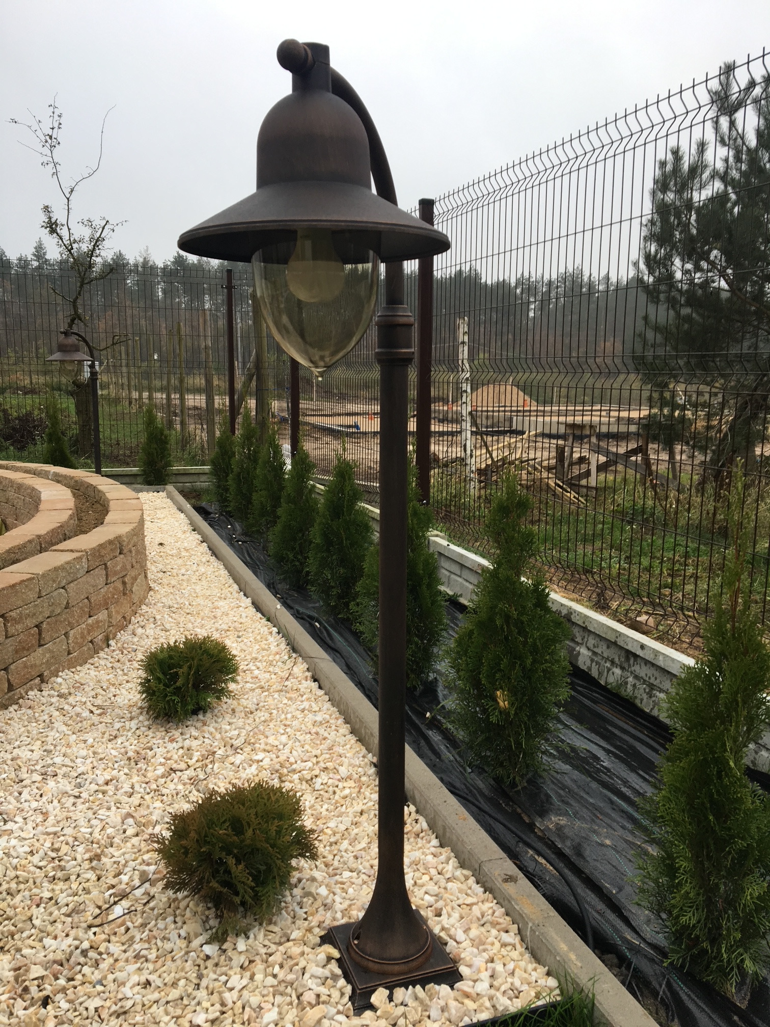 Rustykalna lampa ogrodowa z kloszem w kształcie dzwonu oświetla ścieżkę z białego żwiru, obok posadzone tuje, w tle ażurowe ogrodzenie.