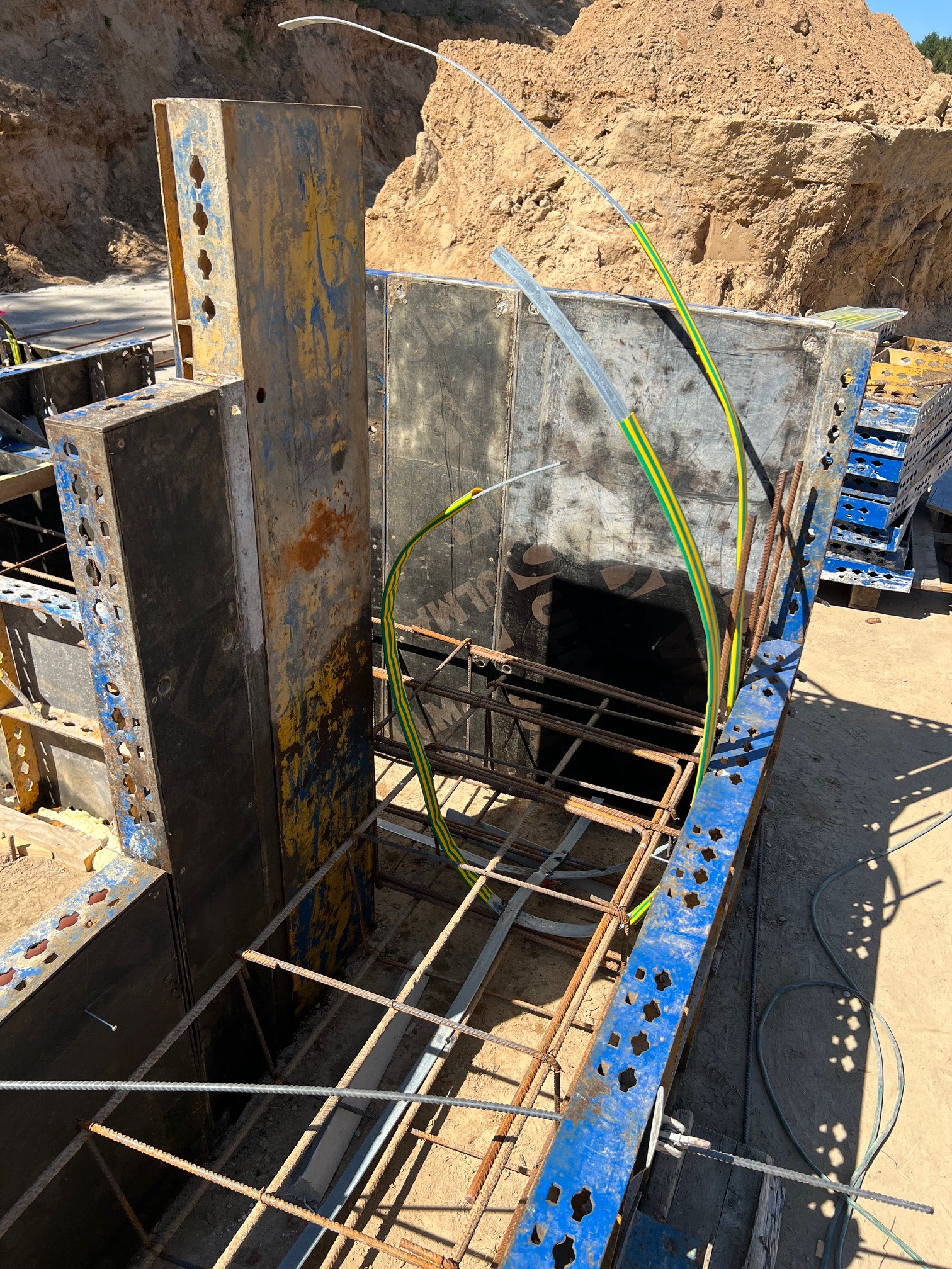 Szczegółowy widok z góry na metalowe szalunki zbrojeniowe z wystającymi przewodami ochronnymi w żółto-zielonej izolacji, przygotowane do zalania betonem na tle wykopu ziemnego.