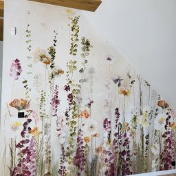 Tapetowanie_Blumal - Ściana w pokoju na poddaszu z drewnianymi belkami, pokryta fototapetą z motywem kwitnących kwiatów w odcieniach fioletu, pomarańczy i bieli, stylizowaną na akwarele.