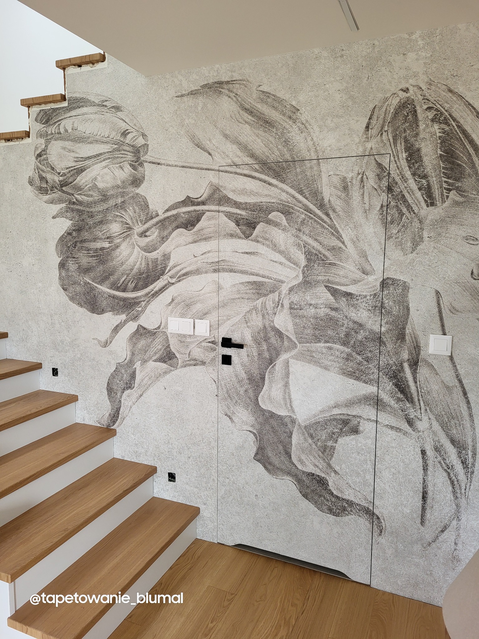 Ściana z fototapetą przedstawiającą duży kwiatowy motyw w odcieniach szarości, z ukrytymi drzwiami i fragmentem schodów z drewnianymi stopniami.
