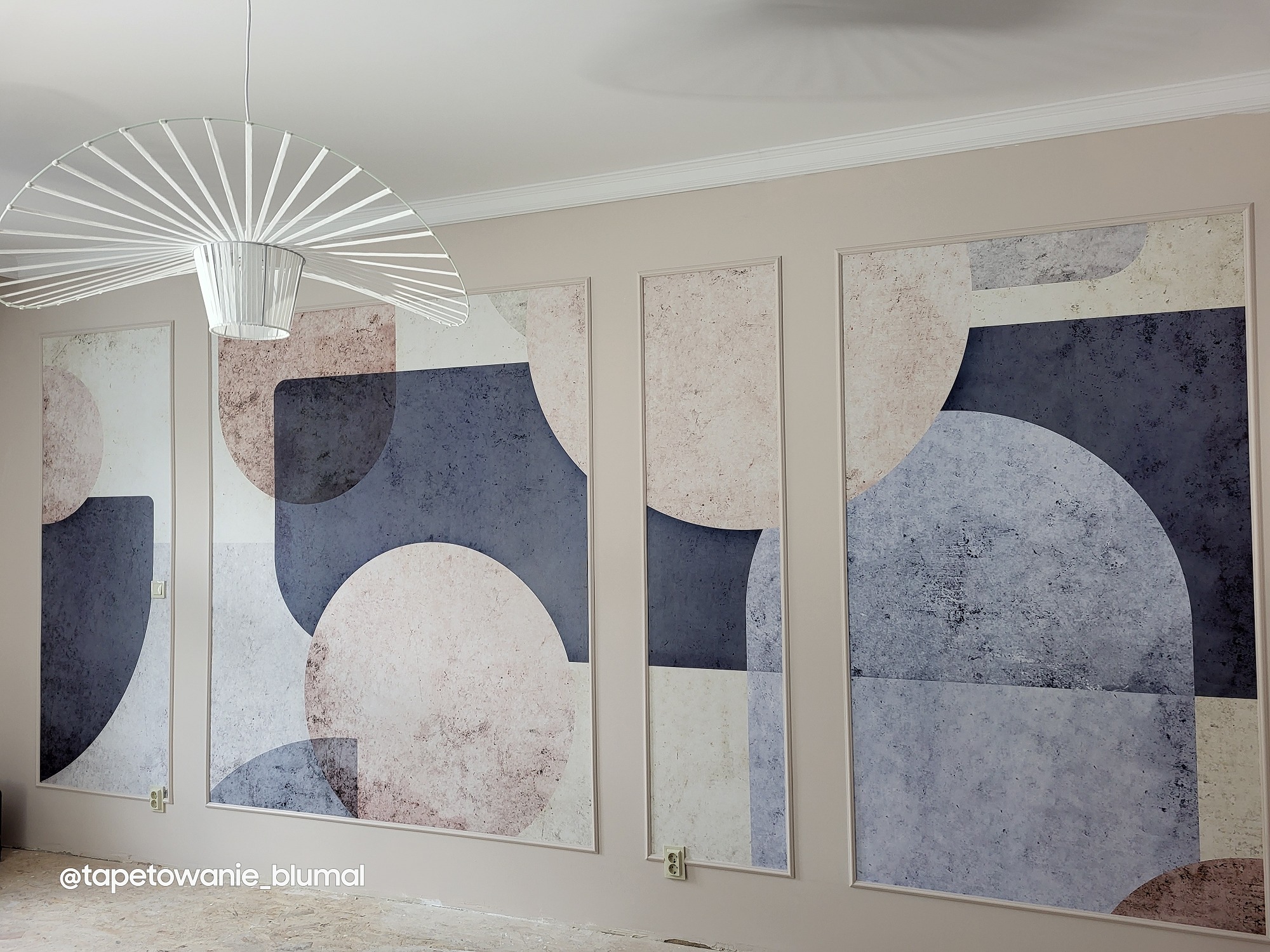 Tapeta w geometryczne wzory (koła i prostokąty) w odcieniach szarości i beżu, zamontowana w panelach ściennych ze sztukaterią, oświetlona przez designerską lampę wiszącą.