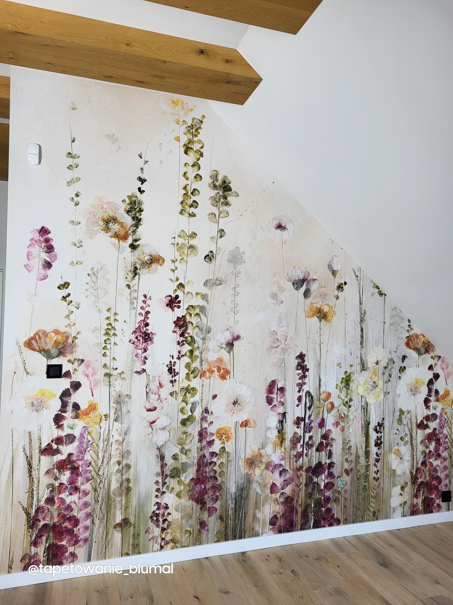 Ściana w pokoju na poddaszu z drewnianymi belkami, pokryta fototapetą z motywem kwitnących kwiatów w odcieniach fioletu, pomarańczy i bieli, stylizowaną na akwarele.