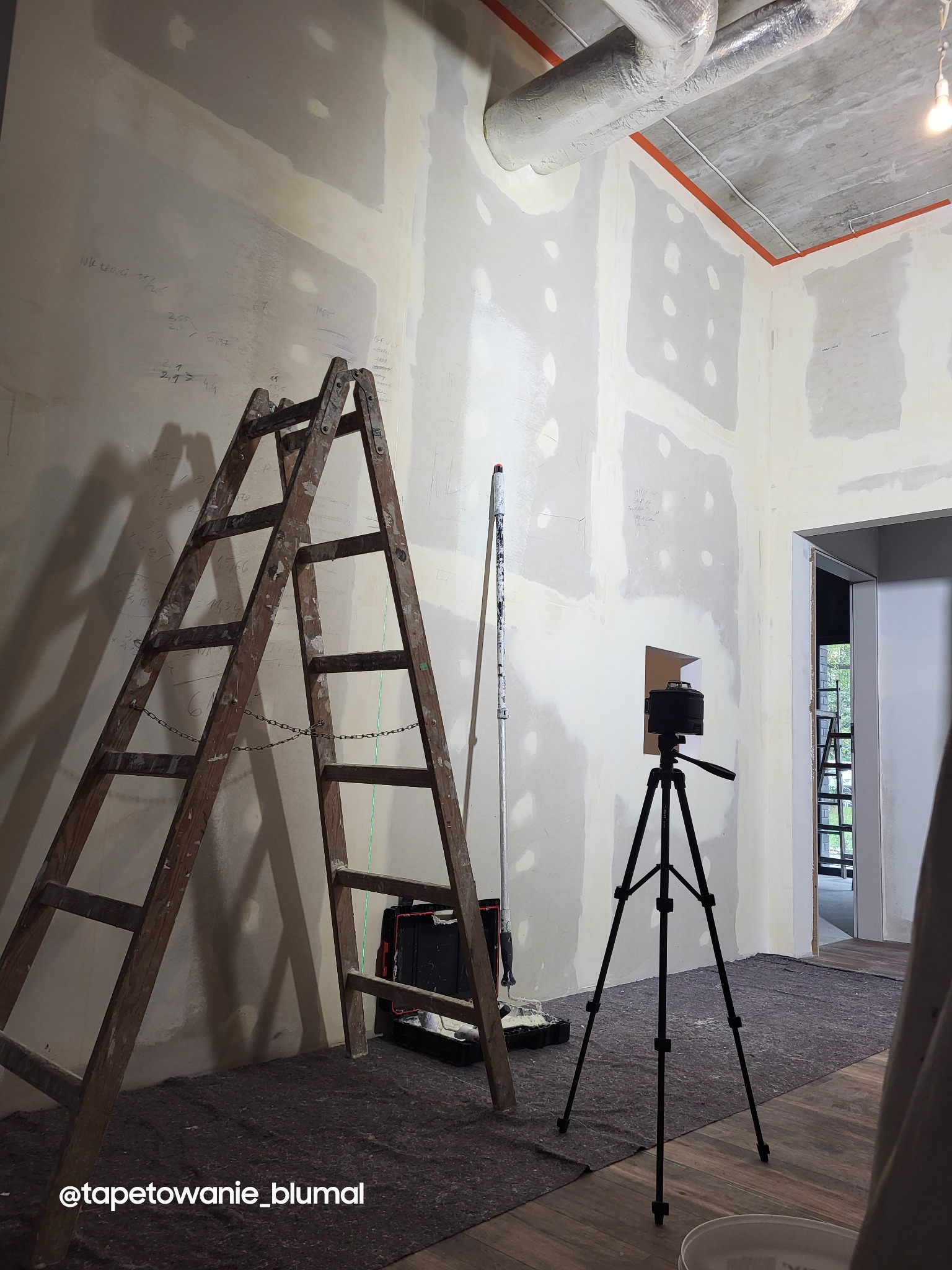 Przygotowanie ścian do tapetowania: drewniana drabina, wałek malarski, niwelator laserowy na trójnogu i ściany z widocznymi śladami szpachlowania i notatkami o wymiarach.