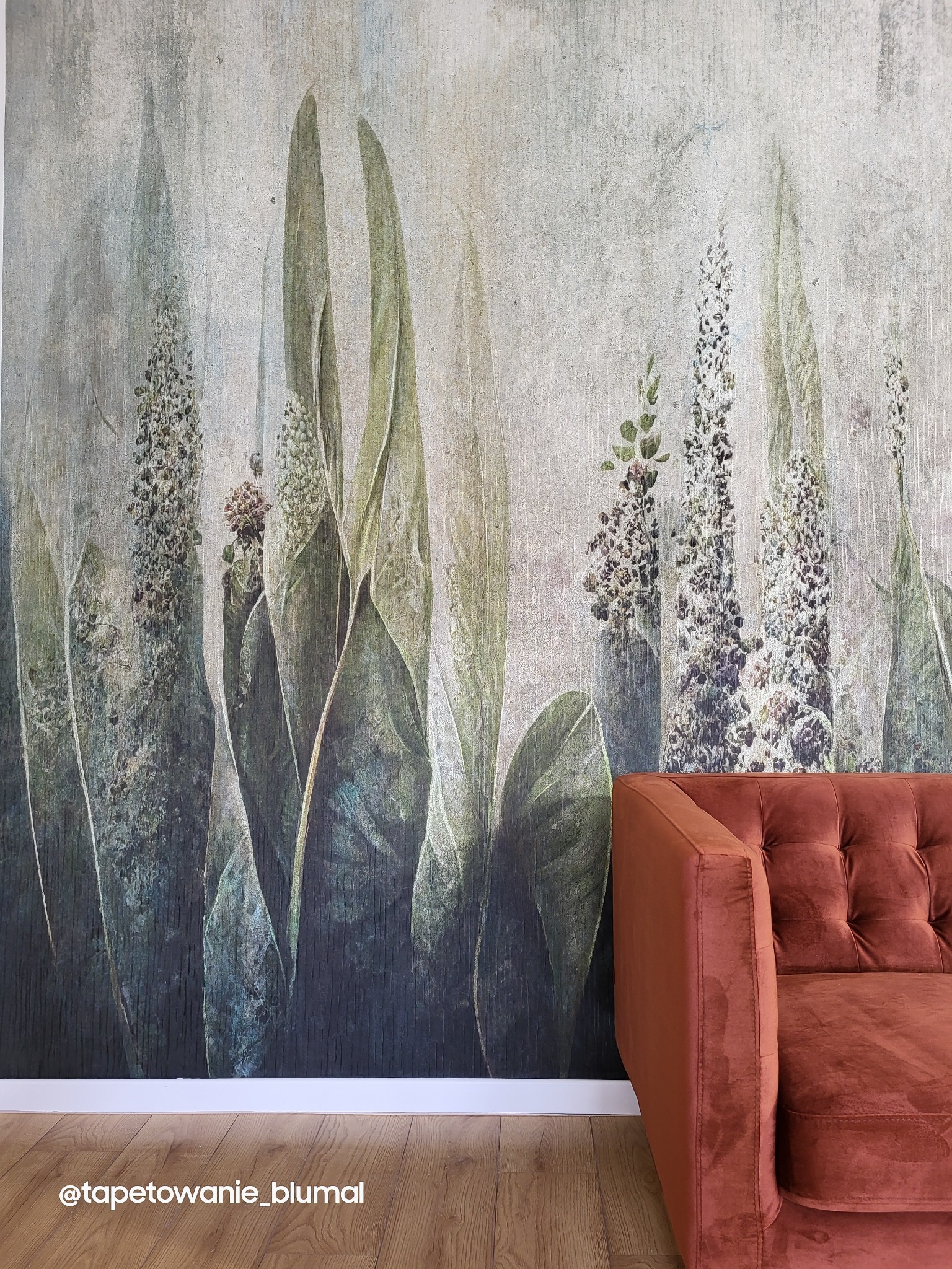 Fototapeta z motywem roślinnym w odcieniach zieleni i szarości, uzupełniona fragmentem bordowej, pikowanej sofy.