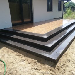 Dam&Bruk - Nowoczesne schody wejściowe z ciemnoszarej kostki brukowej prowadzące na drewniany taras, z fragmentem węża ogrodowego w piasku.