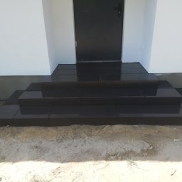 Dam&Bruk - Nowe, czarne, granitowe schody wejściowe do domu, prowadzące do czarnych drzwi, otoczone piaskiem i białą elewacją.