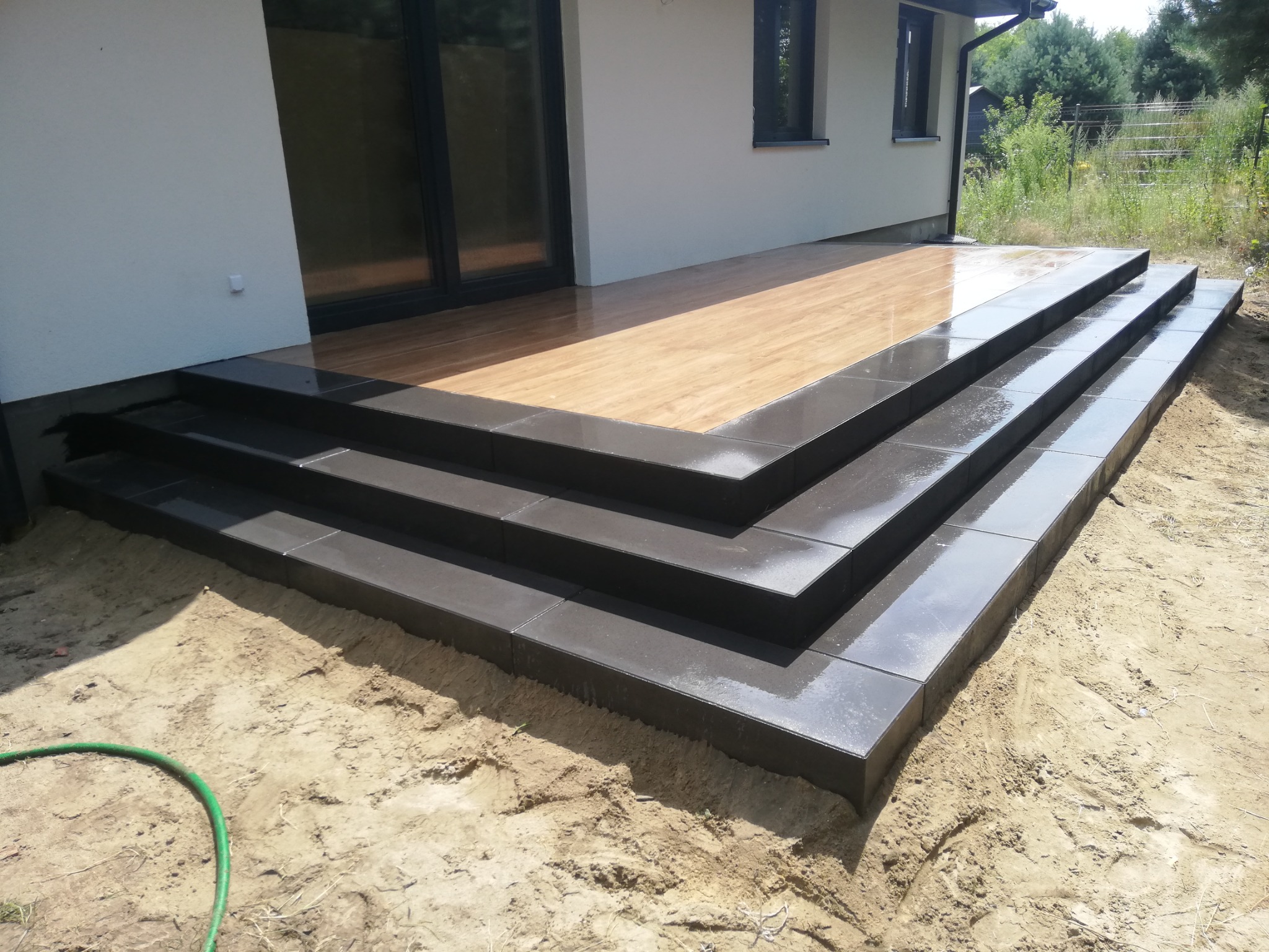 Nowoczesne schody wejściowe z ciemnoszarej kostki brukowej prowadzące na drewniany taras, z fragmentem węża ogrodowego w piasku.