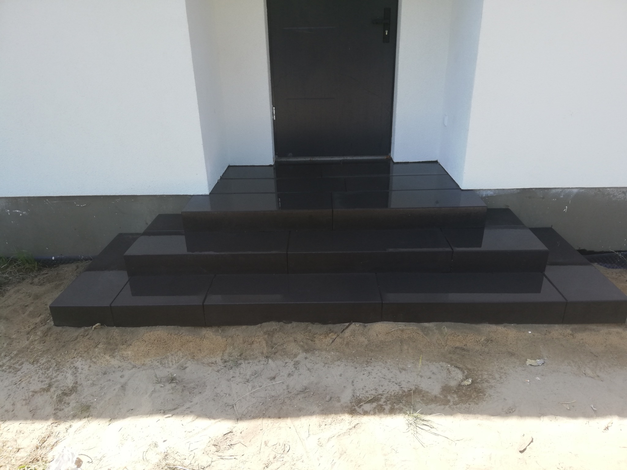 Nowe, czarne, granitowe schody wejściowe do domu, prowadzące do czarnych drzwi, otoczone piaskiem i białą elewacją.