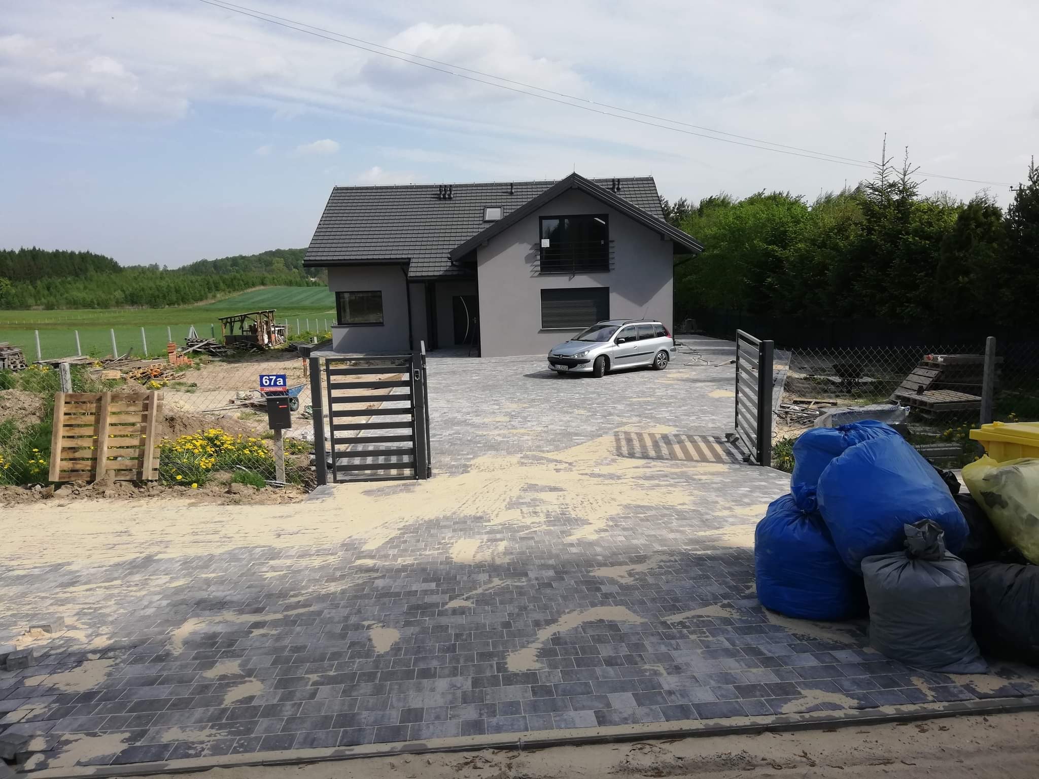 Nowoczesny dom jednorodzinny z szarą elewacją i ciemnym dachem, podjazd wyłożony szarą kostką brukową, widoczne worki z odpadami budowlanymi, teren jeszcze w trakcie prac wykończeniowych.