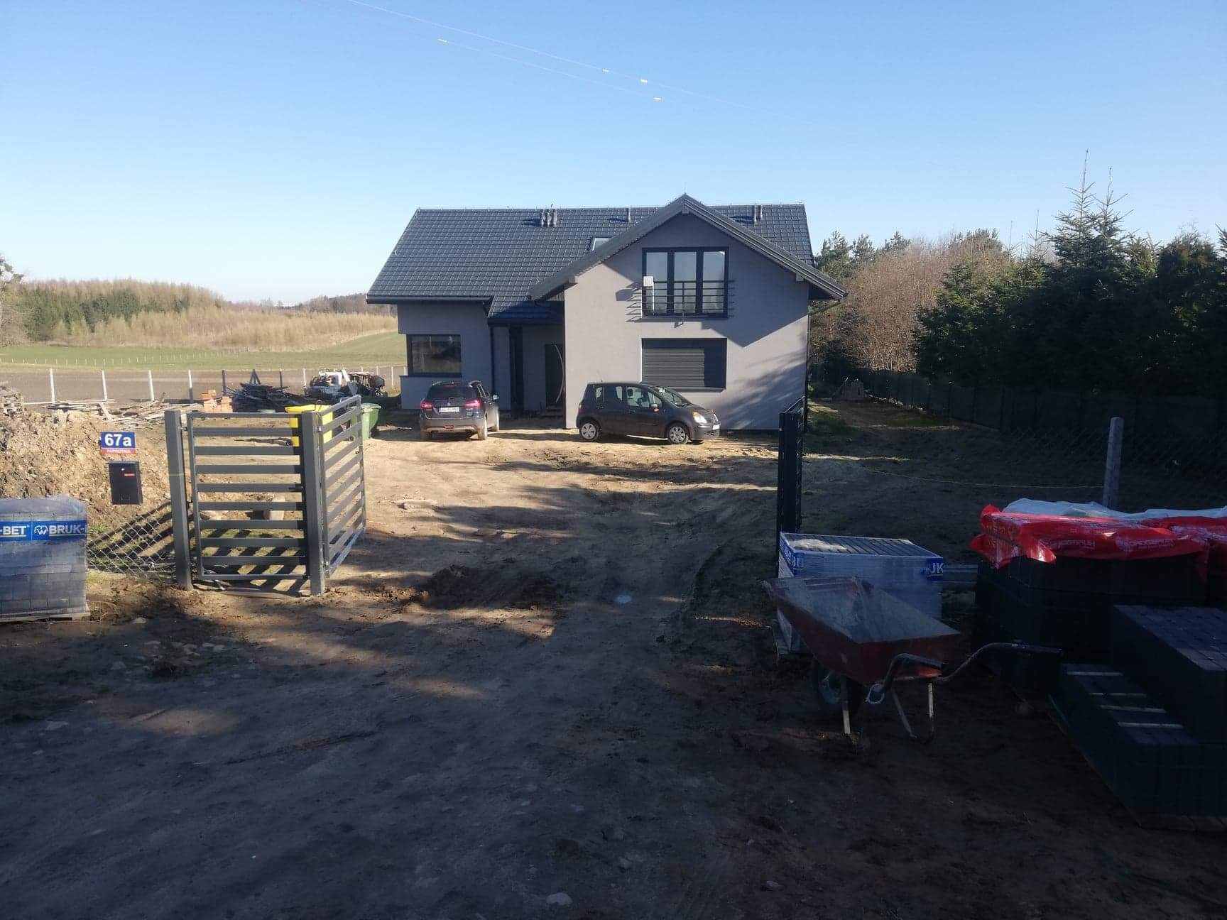 Przygotowany teren pod budowę z widocznym nowym domem, dwoma samochodami, stertą kostki brukowej i taczką, brama wjazdowa zamontowana.