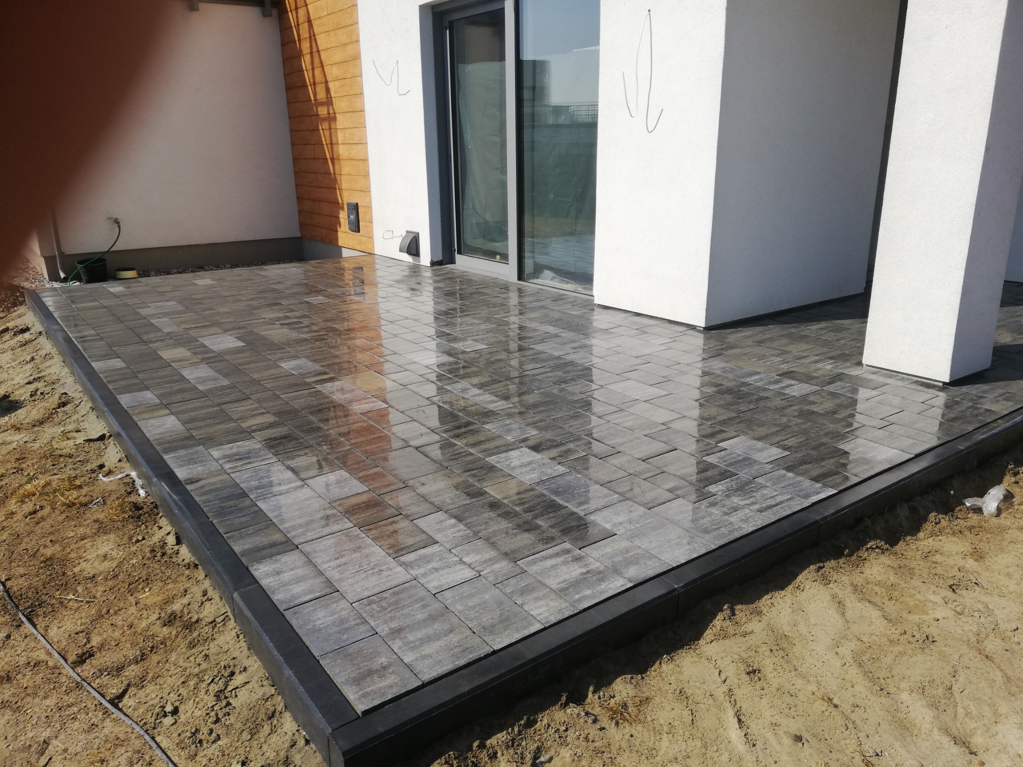 Świeżo ułożona, wilgotna nawierzchnia z kostki brukowej w odcieniach szarości i brązu, obramowana czarnym krawężnikiem, przylegająca do nowoczesnego budynku z przesuwnymi drzwiami tarasowymi.