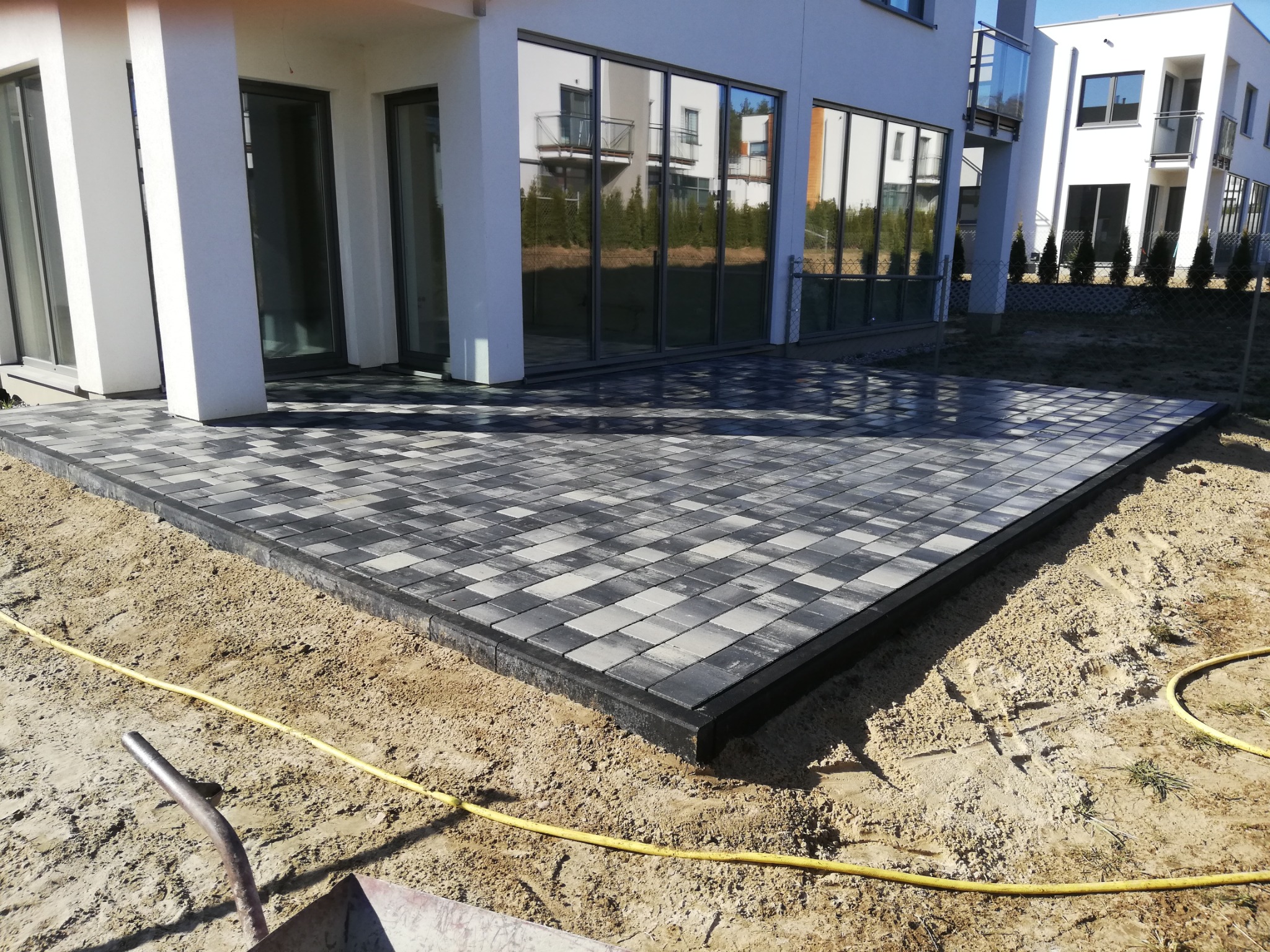 Nowoczesny taras z kostki brukowej w odcieniach szarości, obramowany czarnym obrzeżem, na tle jasnego budynku z dużymi oknami i żółtym wężem do nawadniania leżącym na piasku.
