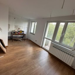 SK-Bud Sebastian Nienajadło - Jasne panele podłogowe w pokoju z oknami i grzejnikiem. Na ścianie widoczne ślady prac remontowych, a na stole narzędzia. Częściowo widoczny karton po panelach.