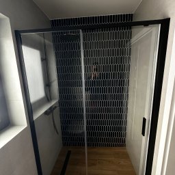 SK-Bud Sebastian Nienajadło - Nowoczesna kabina prysznicowa z czarną ramą i geometrycznymi płytkami w kształcie łezki na ścianie. Drewnopodobna podłoga i minimalistyczny design.