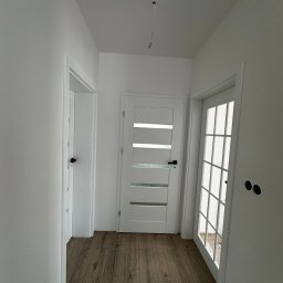 SK-Bud Sebastian Nienajadło - Korytarz po remoncie: drewnopodobne panele, białe ściany i trzy białe drzwi, w tym jedne z przeszkleniami. Widoczne niedokończone gniazdka elektryczne.