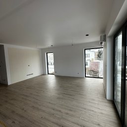 SK-Bud Sebastian Nienajadło - Jasne panele podłogowe w przestronnym, surowym wnętrzu w Jarosławiu. Widoczne przygotowane ściany do malowania i duże okna z czarnymi ramami. Minimalistyczny styl.