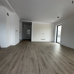 SK-Bud Sebastian Nienajadło - Jasne panele podłogowe w przestronnym, surowym wnętrzu z białymi ścianami i dużym oknem. Widoczne przygotowania do wykończenia, wystające kable.