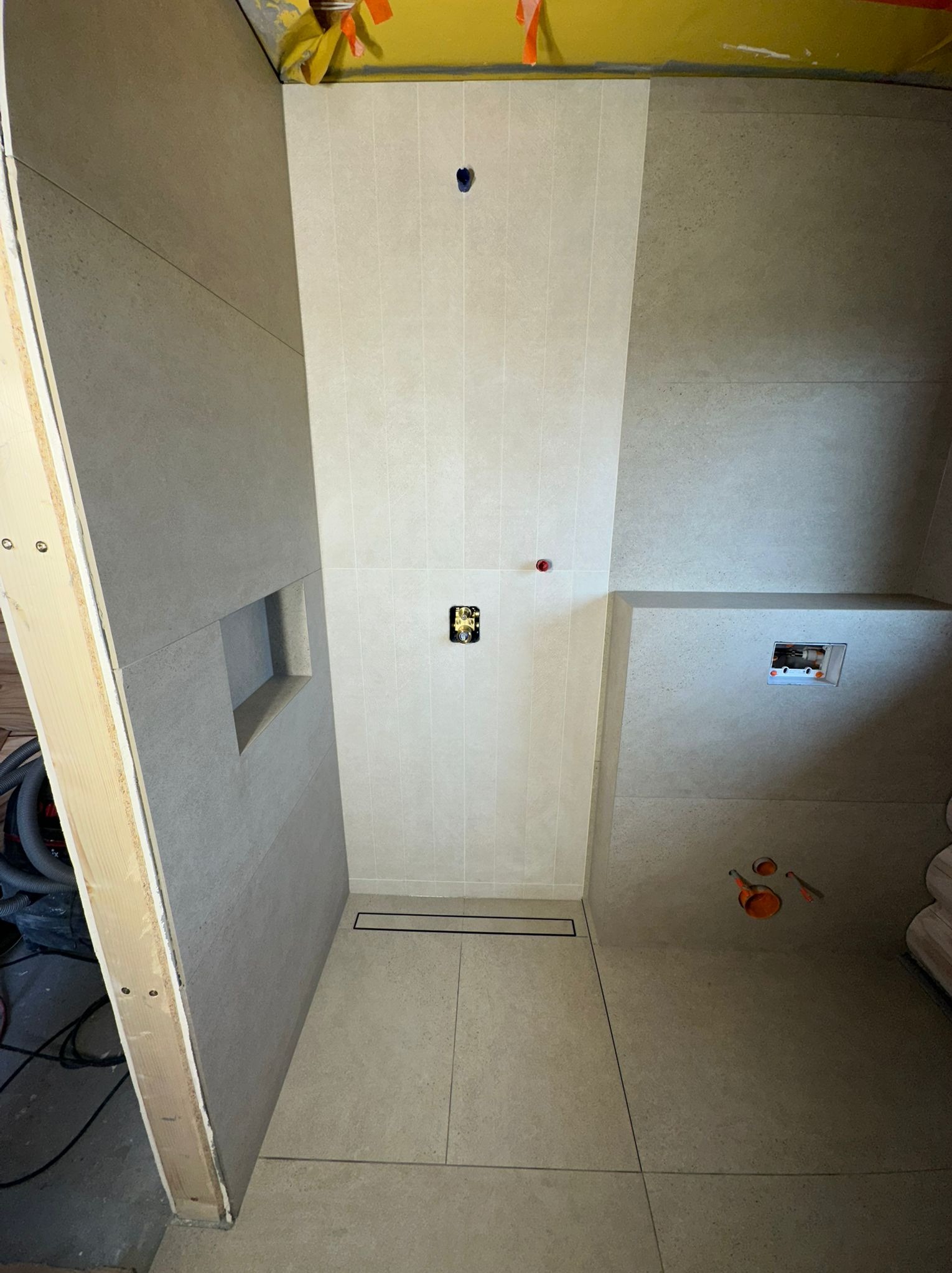 Wykończona kabina prysznicowa z odpływem liniowym i wnęką w ścianie. Widoczne przyłącza hydrauliczne i elektryczne. Surowy stan wykończenia, widoczne zabezpieczenia.