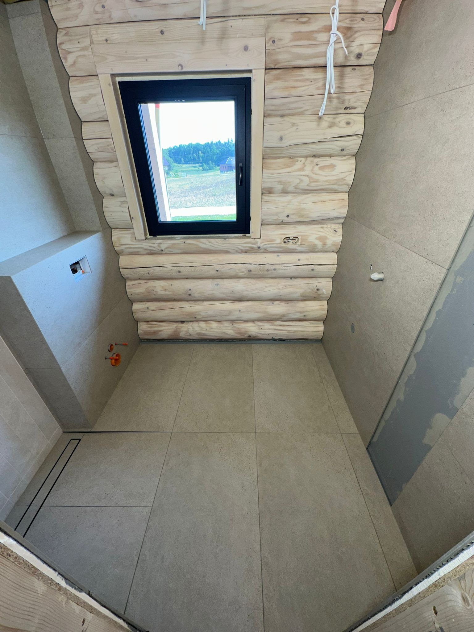 Łazienka w trakcie wykończenia: drewniane ściany, okno z widokiem na zewnątrz, szare płytki na podłodze i ścianach. Widoczne elementy instalacji.