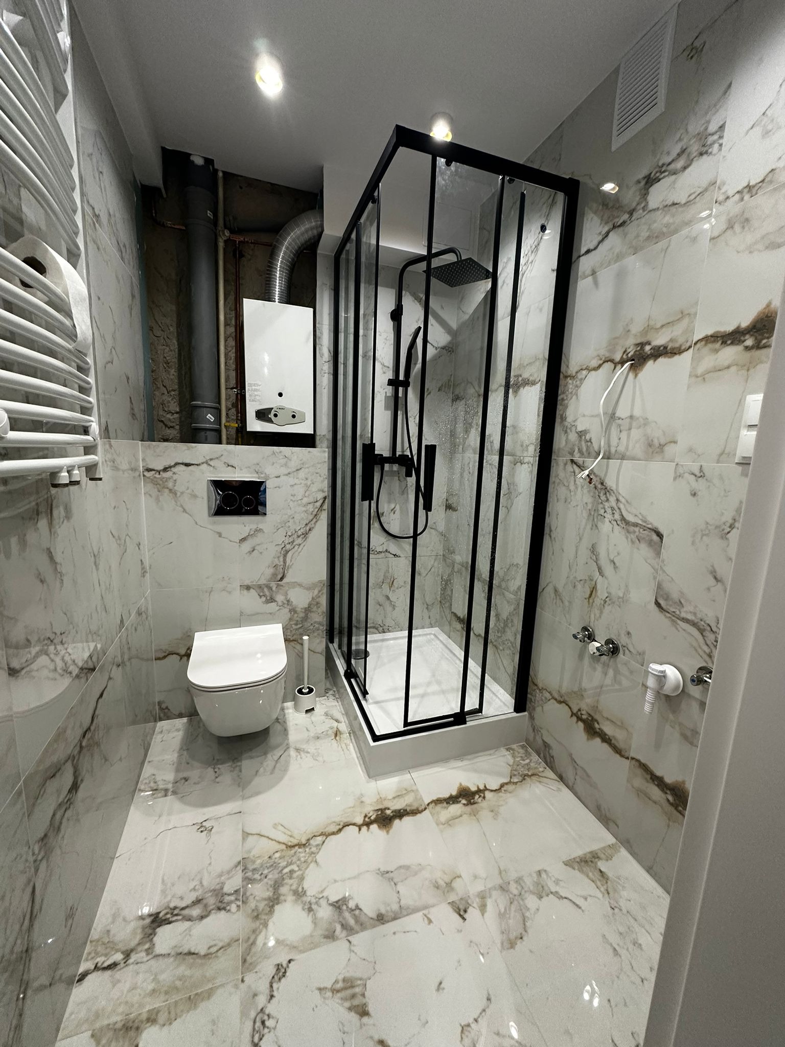 Nowoczesna łazienka z kabiną prysznicową typu walk-in z czarnymi profilami, marmurowe płytki na ścianach i podłodze, wisząca toaleta i grzejnik drabinkowy.