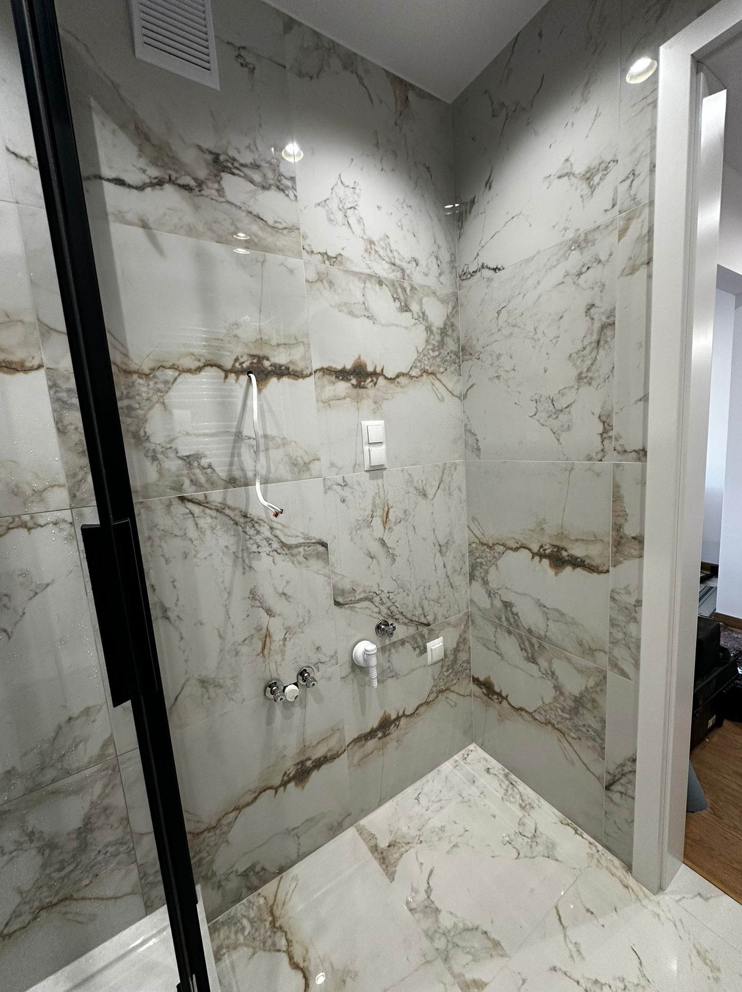 Kabina prysznicowa z marmurowymi płytkami w odcieniach bieli i brązu. Widoczne baterie, czarna rama kabiny i wystające przewody elektryczne. Nowoczesny design, surowe wykończenie.