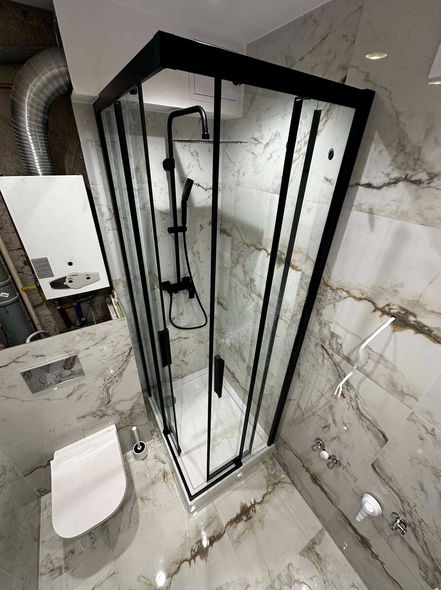 Nowoczesna kabina prysznicowa z czarną ramą i deszczownicą w wykończonej łazience z marmurowymi płytkami. Widoczny podtynkowy system WC i piec gazowy.