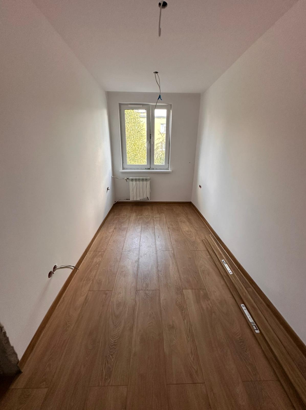 Jasne panele podłogowe w pokoju po remoncie. Świeżo pomalowane ściany, widoczne kable elektryczne i grzejnik pod oknem. Częściowo ułożone listwy przypodłogowe.