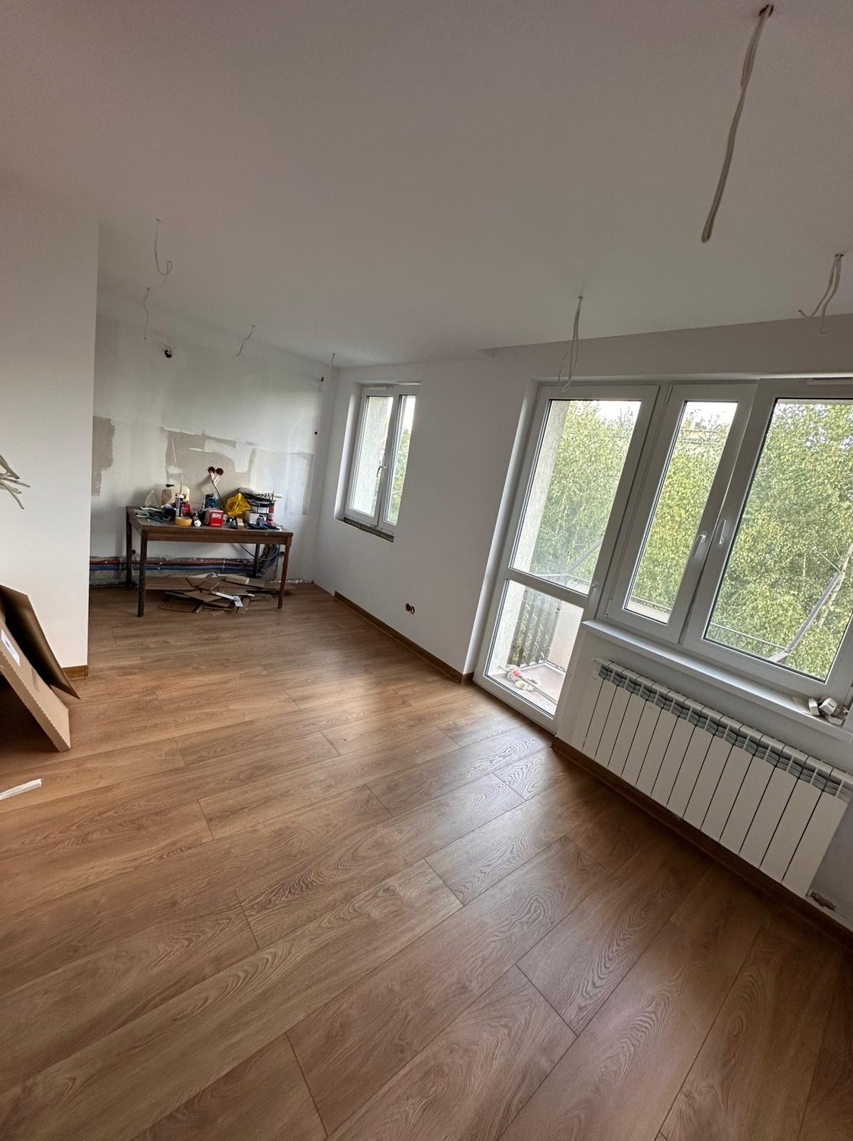 Jasne panele podłogowe w pokoju z oknami i grzejnikiem. Na ścianie widoczne ślady prac remontowych, a na stole narzędzia. Częściowo widoczny karton po panelach.