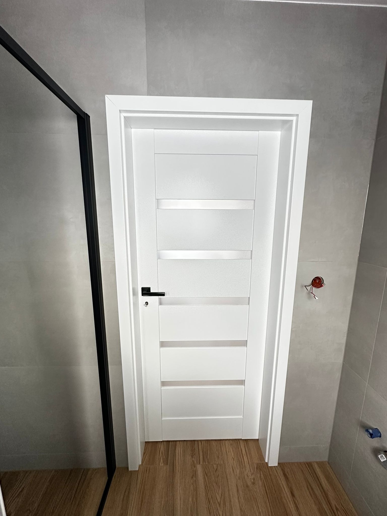 Białe, nowoczesne drzwi wewnętrzne z przeszkleniami w łazience wykończonej szarą płytką. Widoczny fragment kabiny prysznicowej z czarną ramą i drewnopodobna podłoga.