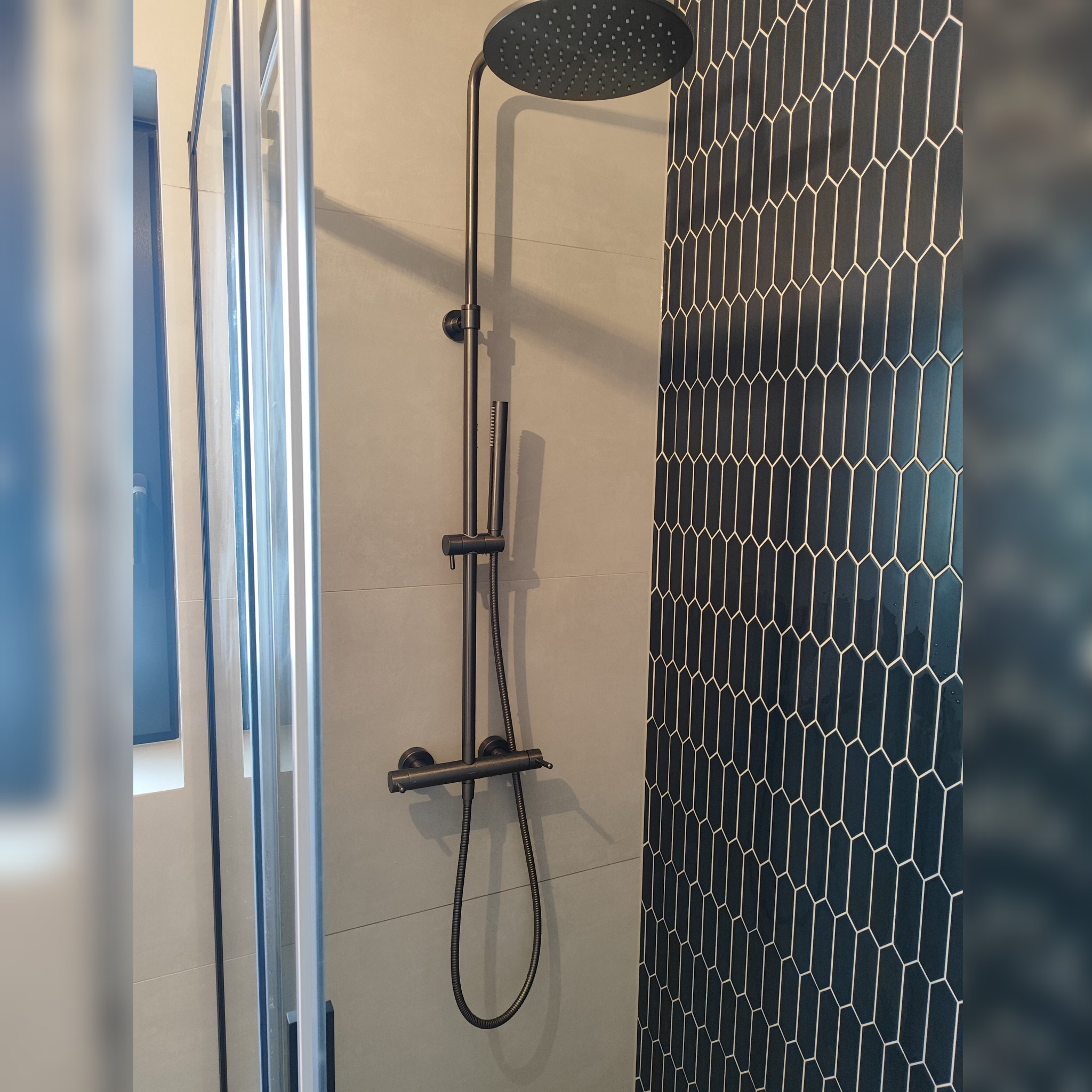 Nowoczesna kabina prysznicowa z czarną armaturą i geometrycznymi płytkami w odcieniach granatu. Minimalistyczny design, szklane drzwi, stonowana kolorystyka.