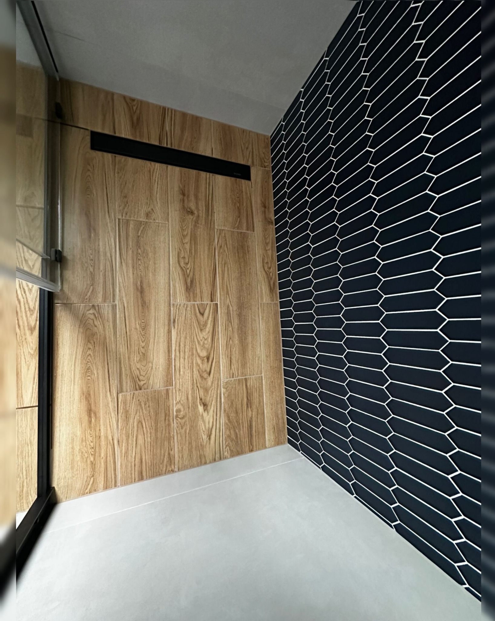 Nowoczesna łazienka z drewnopodobnymi płytkami i czarną ścianą z heksagonalnym wzorem. Minimalistyczny design, widoczny odpływ liniowy. Wykończenie z dbałością o detale.