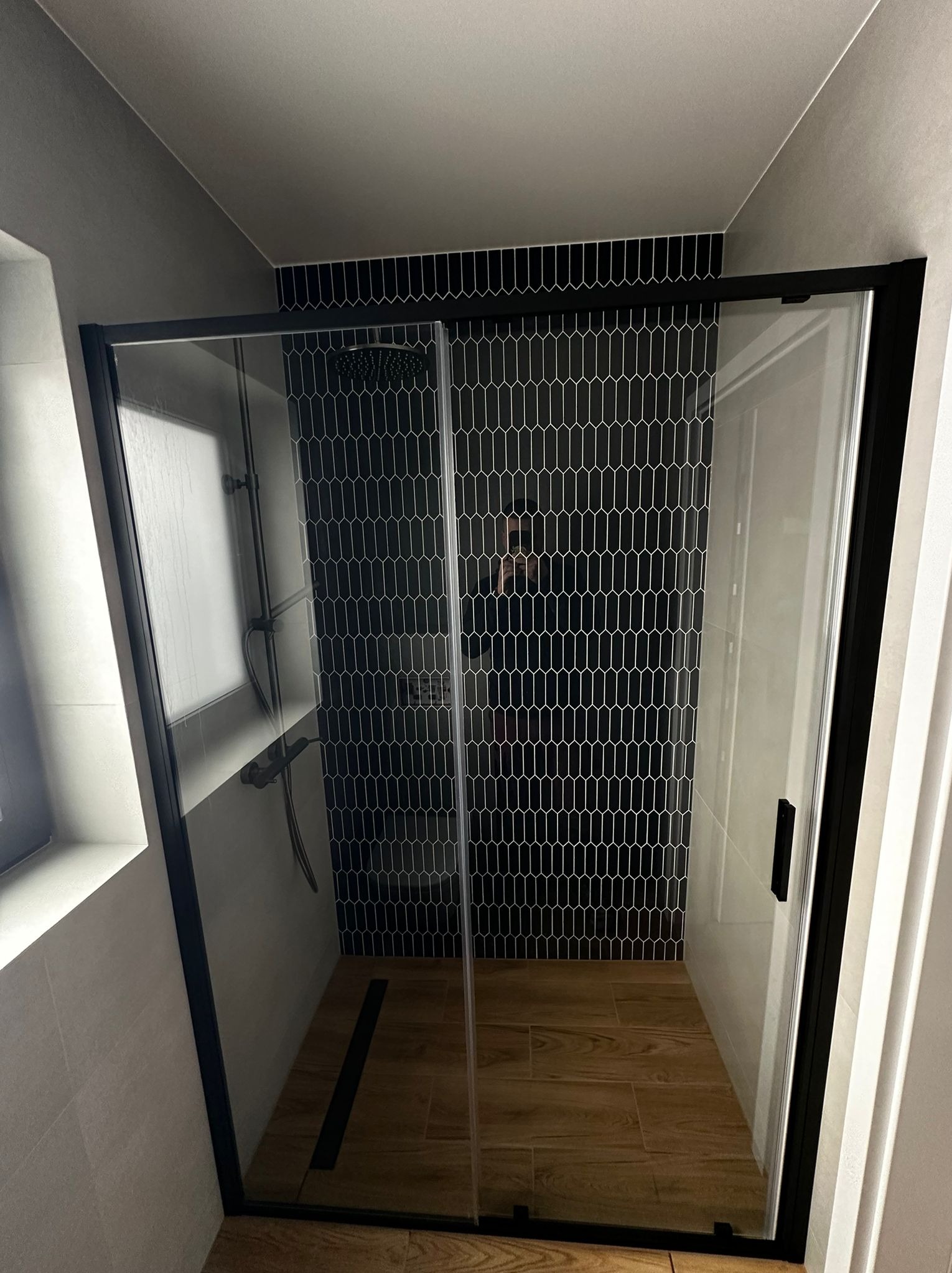 Nowoczesna kabina prysznicowa z czarną ramą i geometrycznymi płytkami w kształcie łezki na ścianie. Drewnopodobna podłoga i minimalistyczny design.