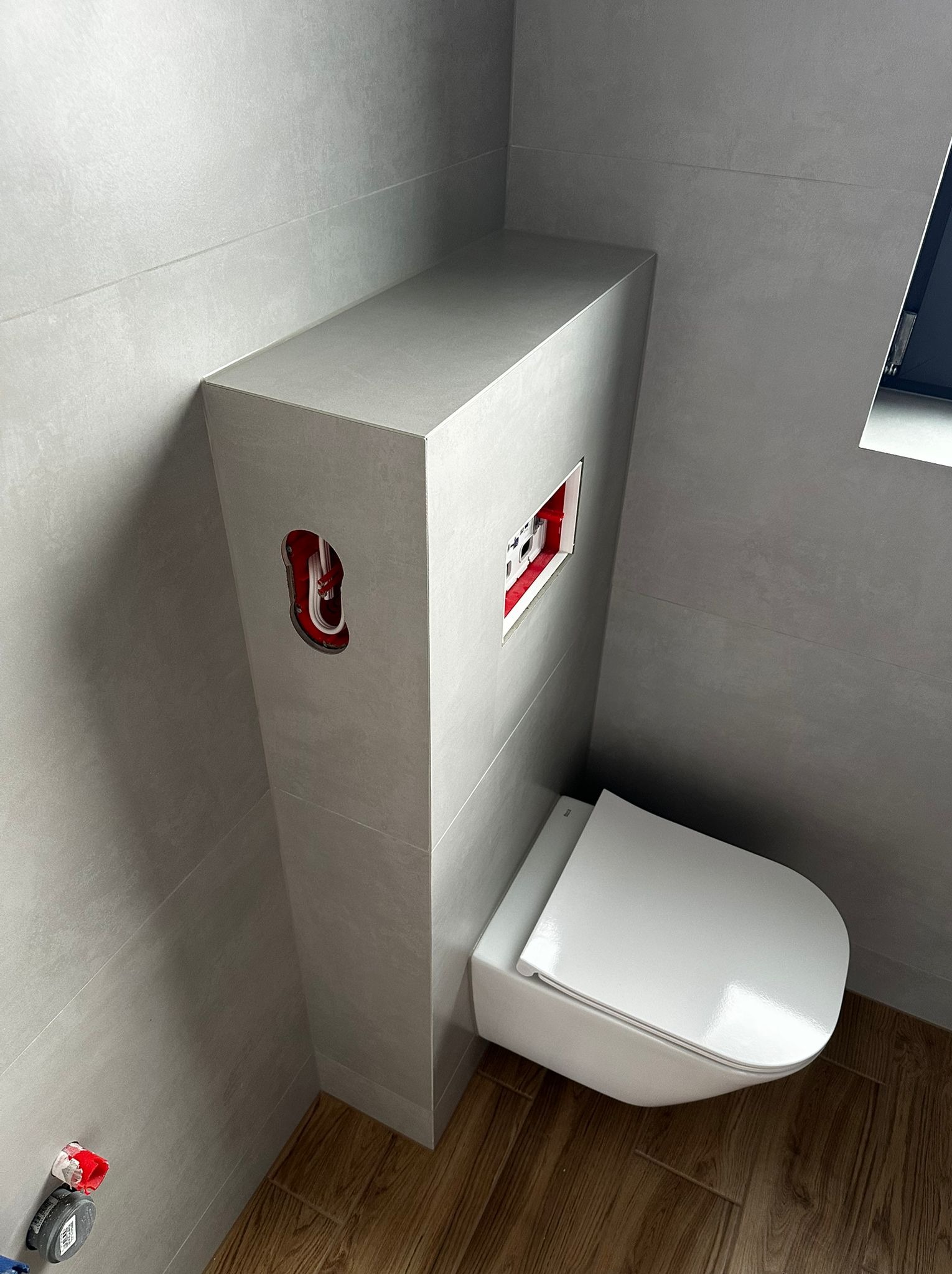 Nowoczesna, podwieszana toaleta w łazience wykończonej szarymi płytkami. Widoczne instalacje w ścianie i drewnopodobna podłoga. Minimalistyczny design.