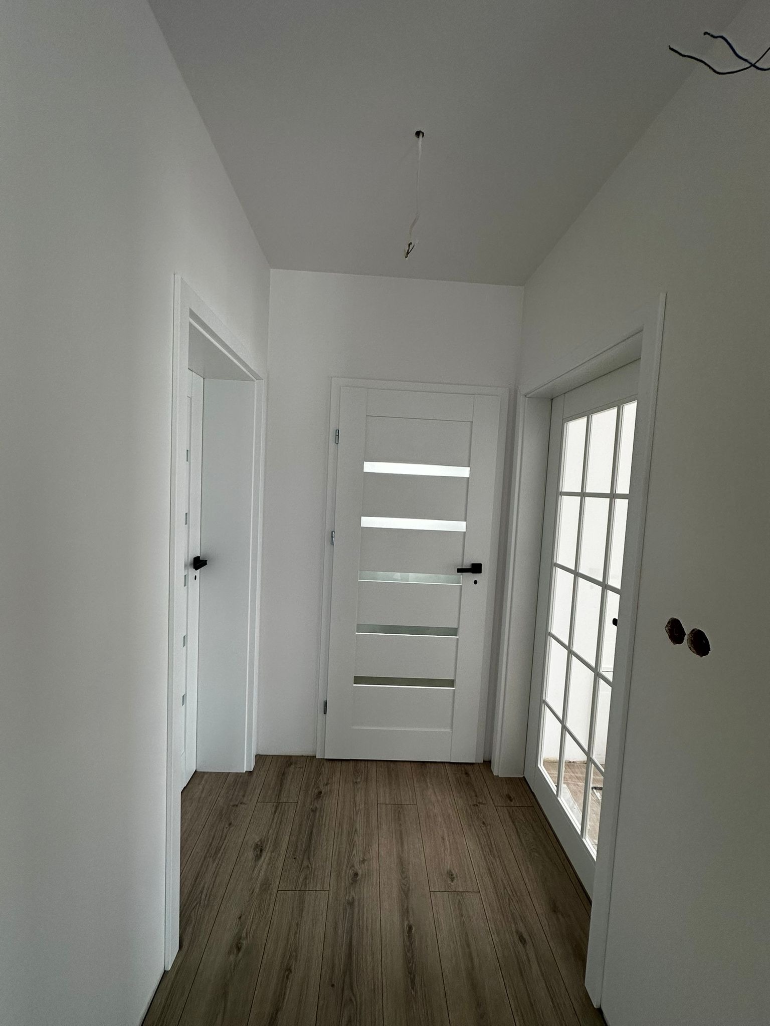 Korytarz po remoncie: drewnopodobne panele, białe ściany i trzy białe drzwi, w tym jedne z przeszkleniami. Widoczne niedokończone gniazdka elektryczne.